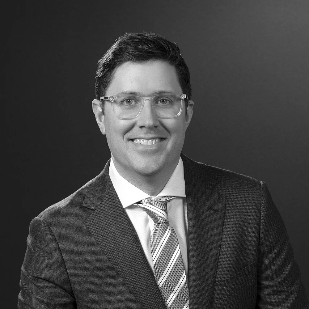 Dr Jack Crozier - Black and white portrait of a man in a suit with glasses, smiling, in front of a dark background.