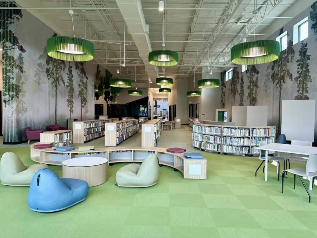 Bright, modern library interior with green ceiling lights, patterned walls, curved bookshelves filled with books, colorful seating, and natural light from high windows.