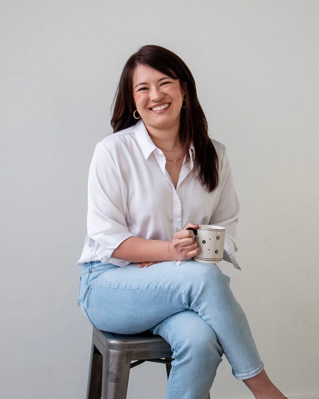 Woman with brown hair wearing a white shirt and light blue jeans, smiling while sitting on a stool and holding a mug against a plain white background.