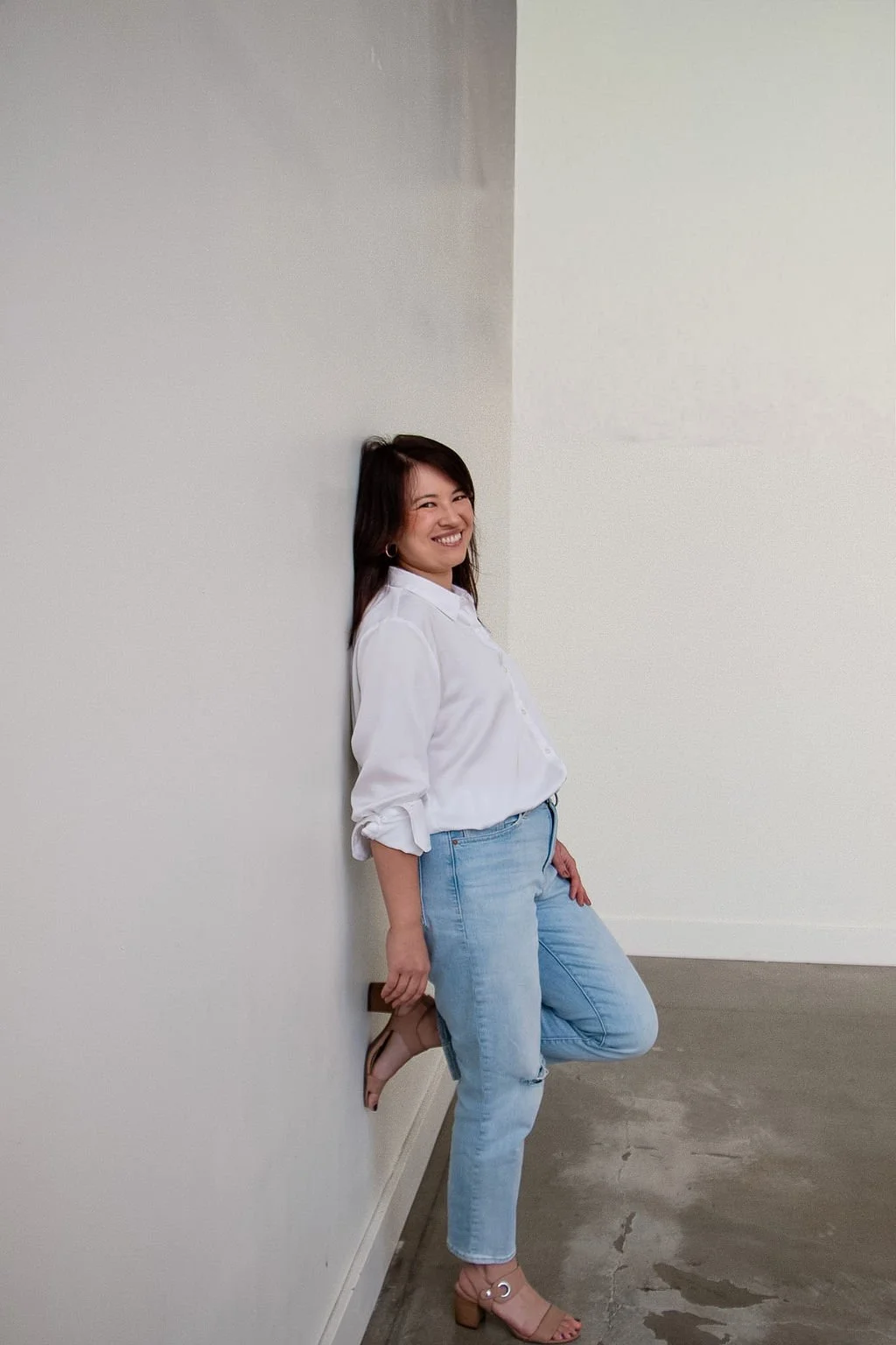 A woman with dark hair and earrings standing against a plain white wall, smiling, wearing a white blouse, light blue jeans, and tan heels.