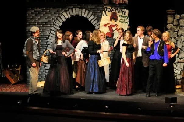 A group of actors performing on stage dressed in period costumes, with a brick archway and a sign reading 'Rosinette' in the background.