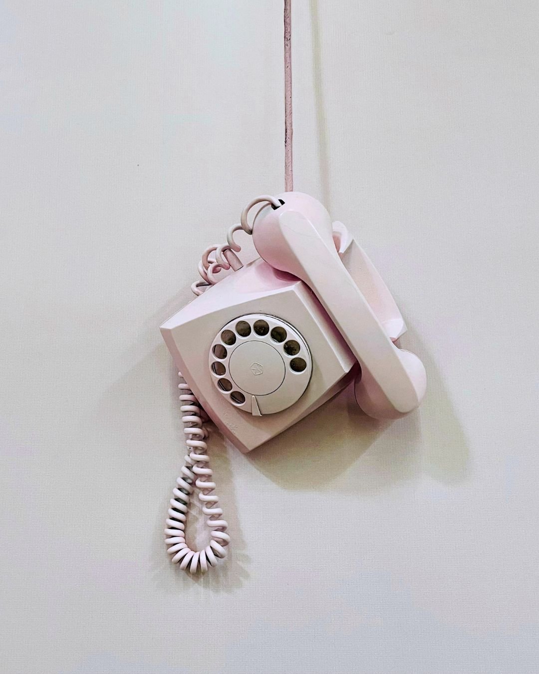 Pink rotary wall phone hanging from the cord against a plain white wall.