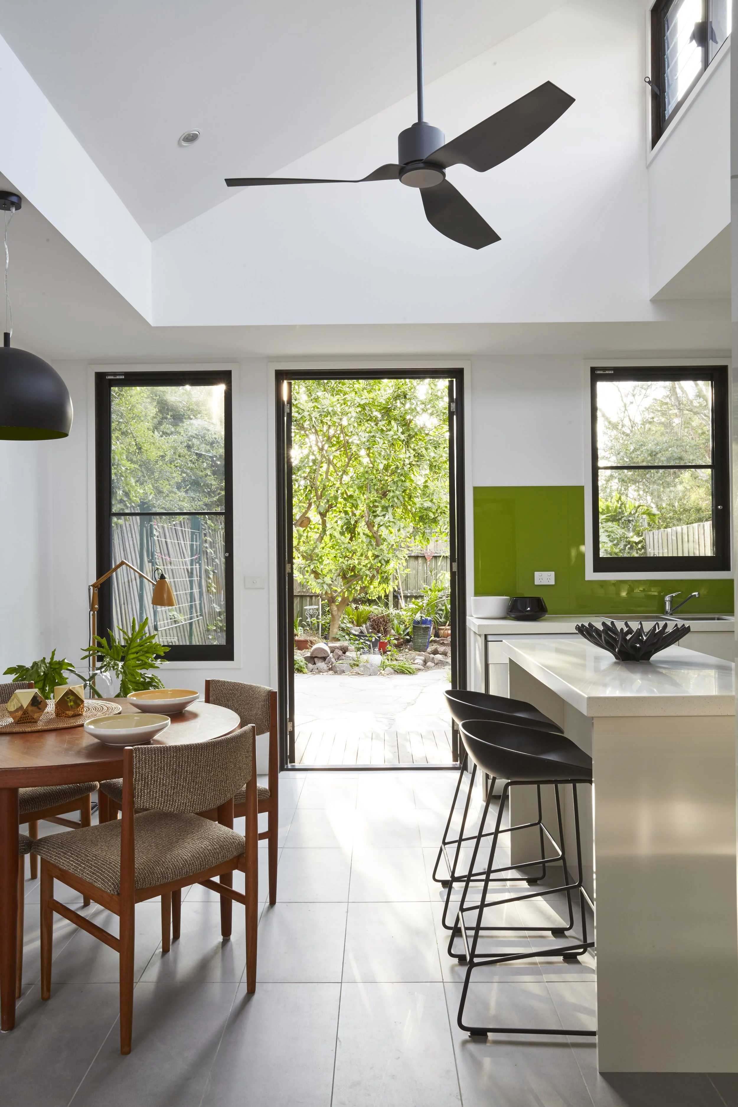 Modern kitchen dining area with a view to a lush garden, featuring a round wooden table, four chairs, black barstools, a green backsplash, a ceiling fan, and large windows.