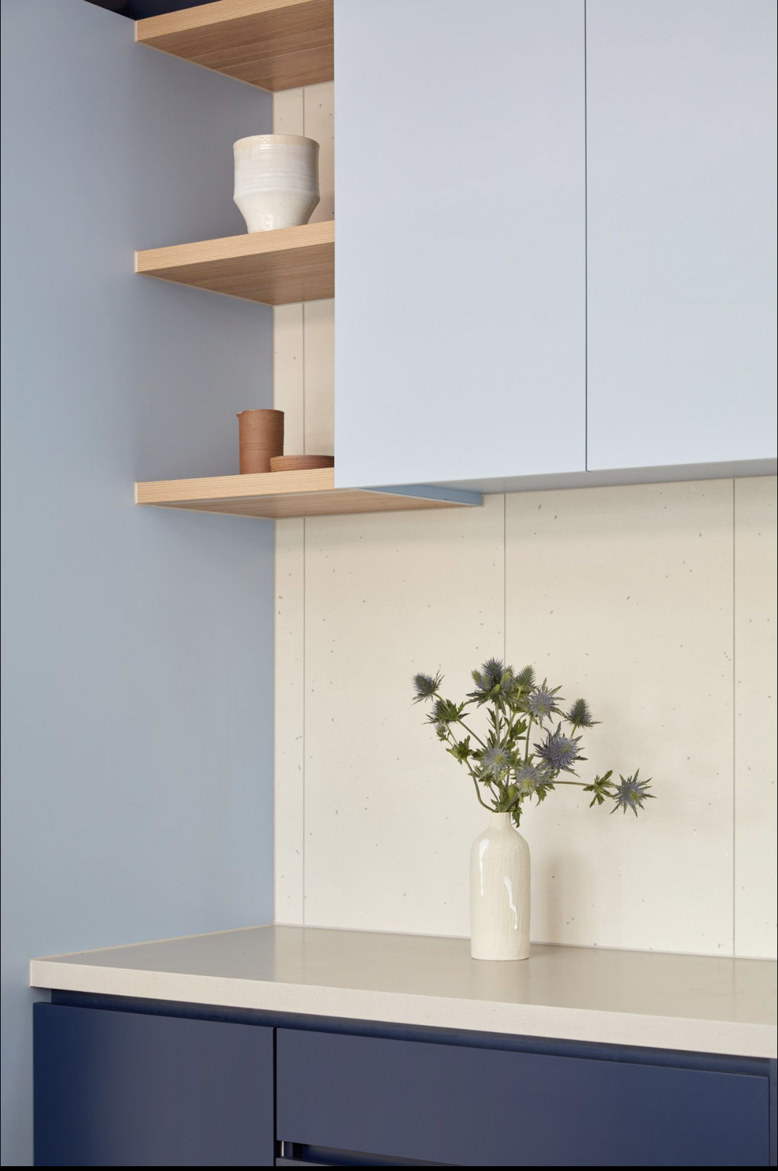 A modern kitchen with light blue cabinets, white countertop, open wooden shelves, and a white vase with purple flowers.