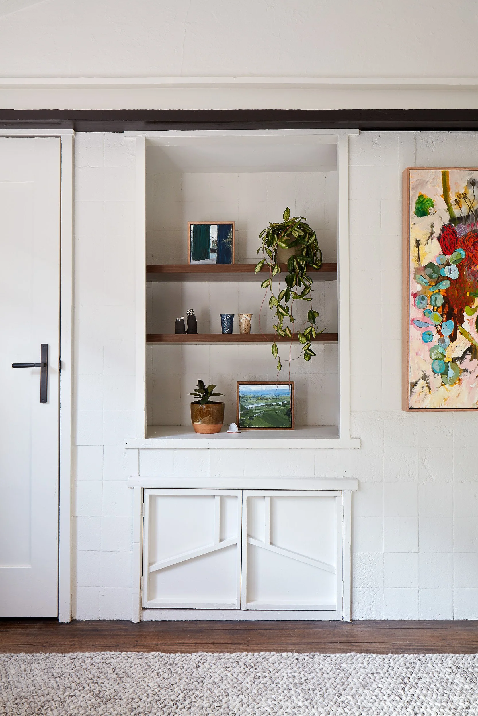 White built-in wall shelf with three wooden shelves holding decorative items, including small pottery, framed artwork, and greenery, next to a colorful abstract painting on the wall.