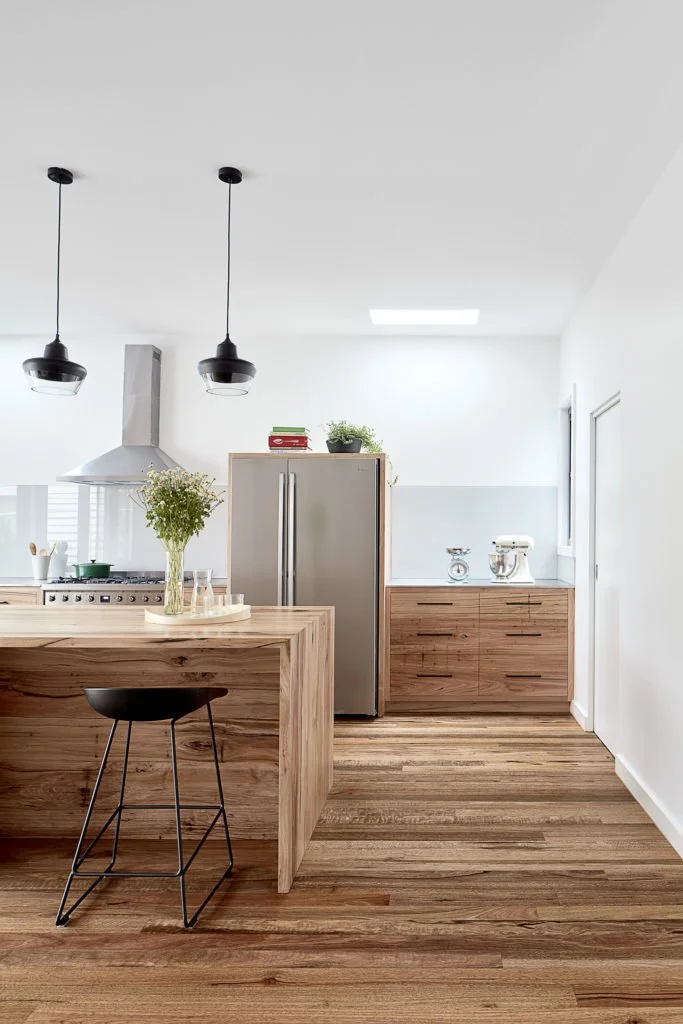 Modern kitchen with wooden island, two black pendant lights, stainless steel refrigerator, wooden cabinets, white walls, and hardwood floors.