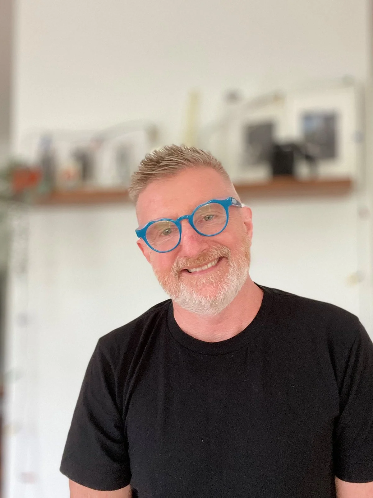 A smiling man with light skin, glasses with blue frames, short gray hair, and a beard, wearing a black t-shirt, standing indoors with a blurred background of a kitchen.