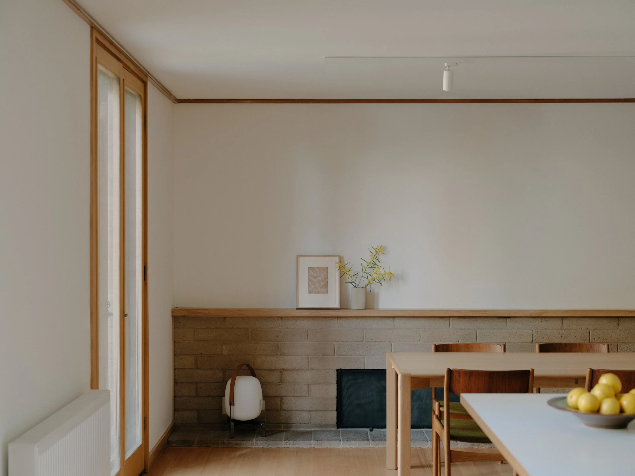 Minimalist dining area with a wooden table and chairs, a white wall with a small art piece and a vase with yellow flowers, a brick fireplace, and a door with glass panels.