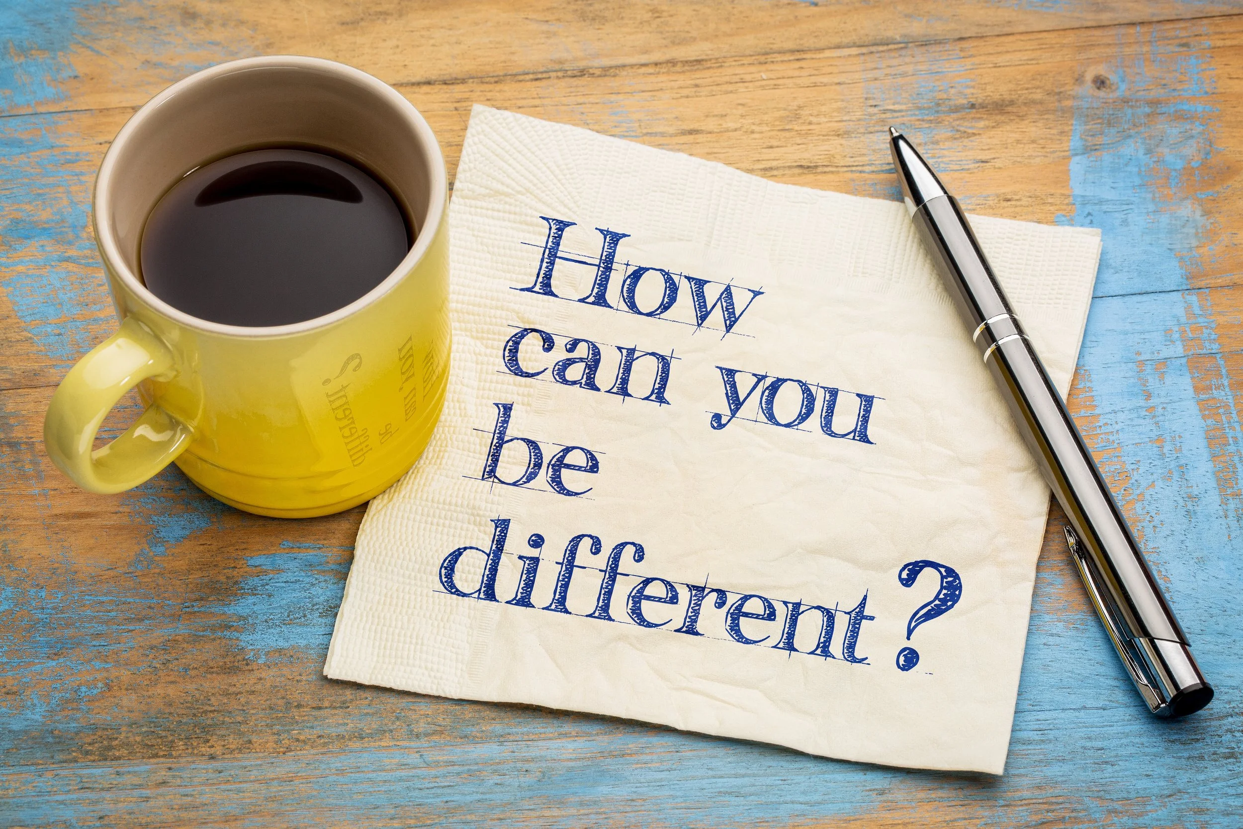 A yellow coffee mug with black coffee, a crumpled napkin with the question 'How can you be different?' written in blue ink, and a black pen on a rustic wooden table with blue paint patches.