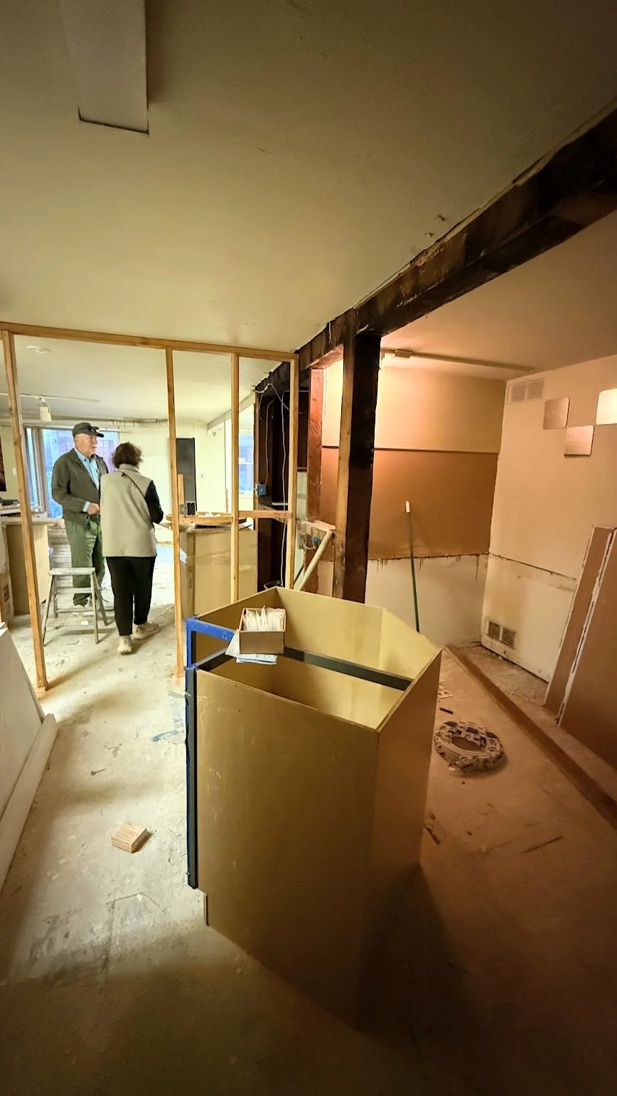 Interior of a building under renovation with exposed wooden framing and construction materials, five people discussing, and construction tools scattered on the floor.