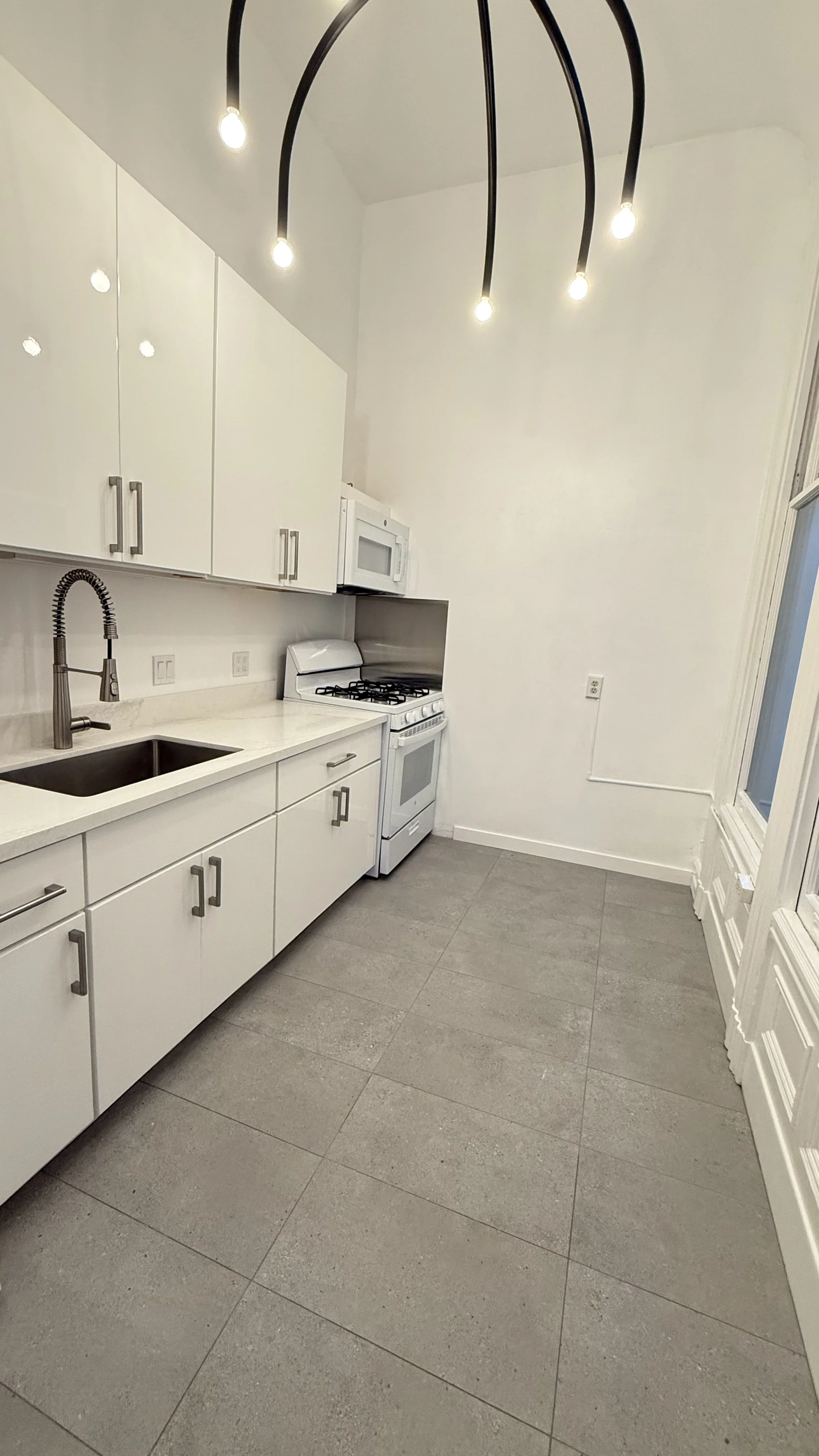 Empty kitchen with white cabinets, stainless steel sink and faucet, gas stove, microwave above stove, and large window with white trim, illuminated by modern ceiling light fixture with five hanging bulbs.