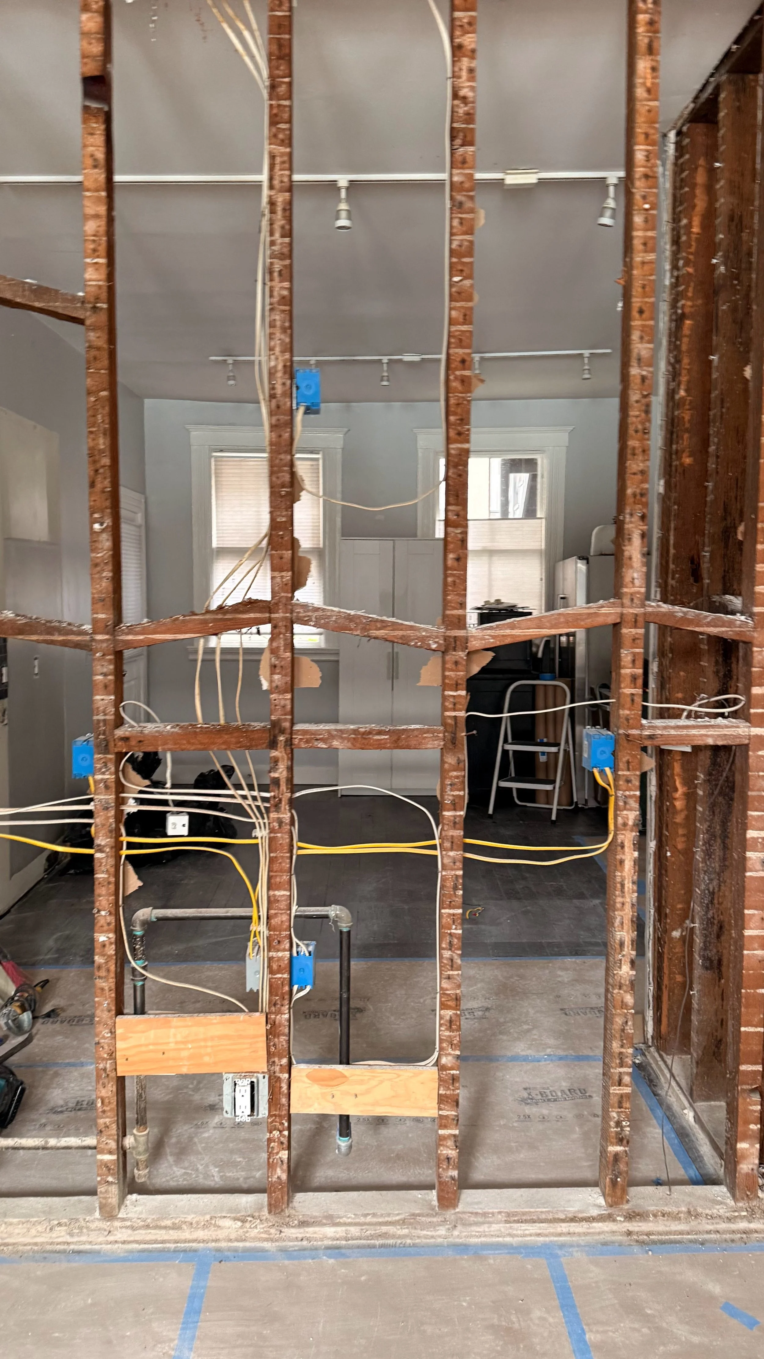 Interior view of a room under construction with exposed wooden framing, electrical wiring, and plumbing visible, and a kitchen area in the background with windows and cabinets.