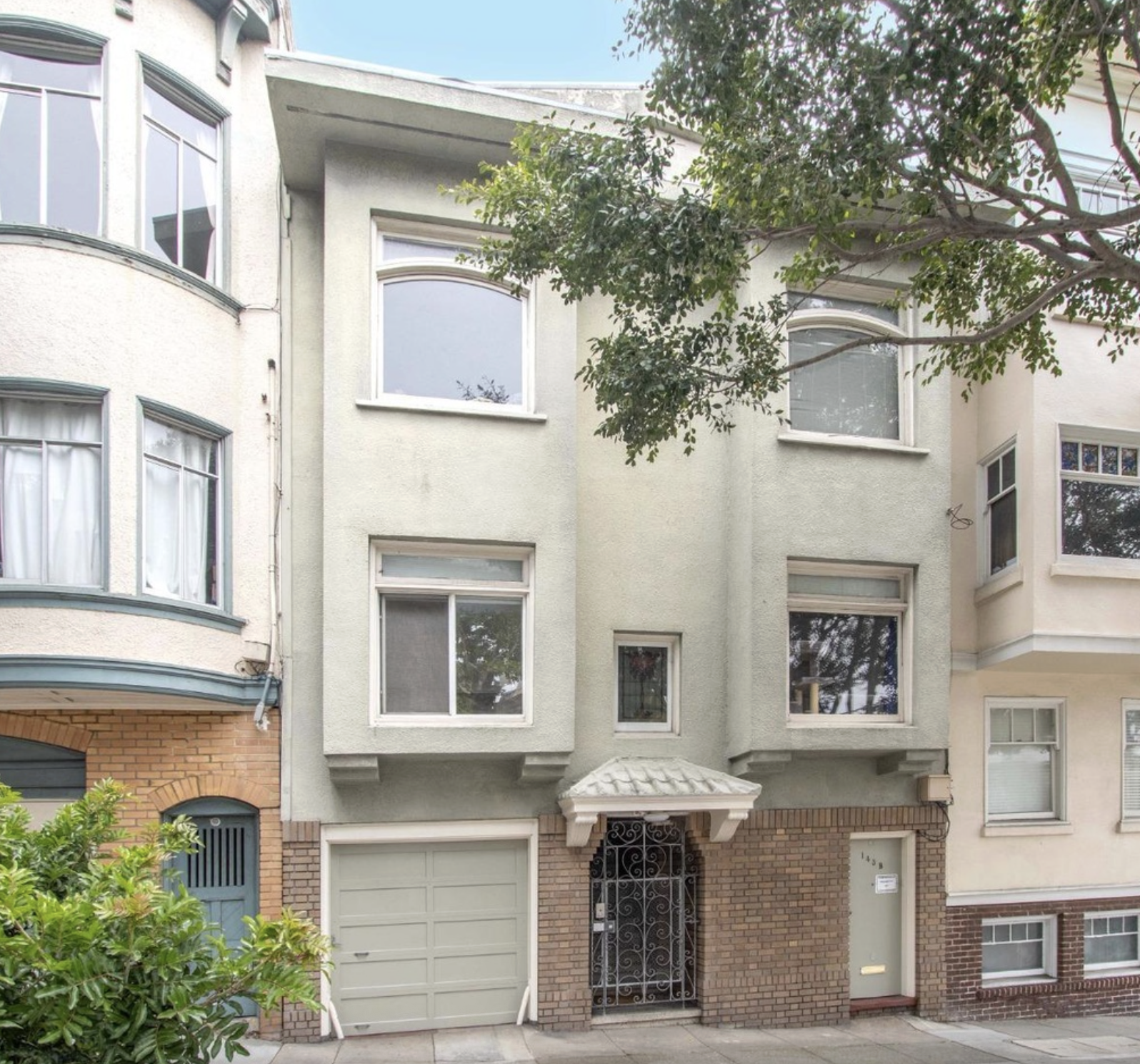 Multi-story residential building with a brick and stucco facade, decorative wrought iron gate, garage door, and multiple rectangular and arched windows, with a tree in front.
