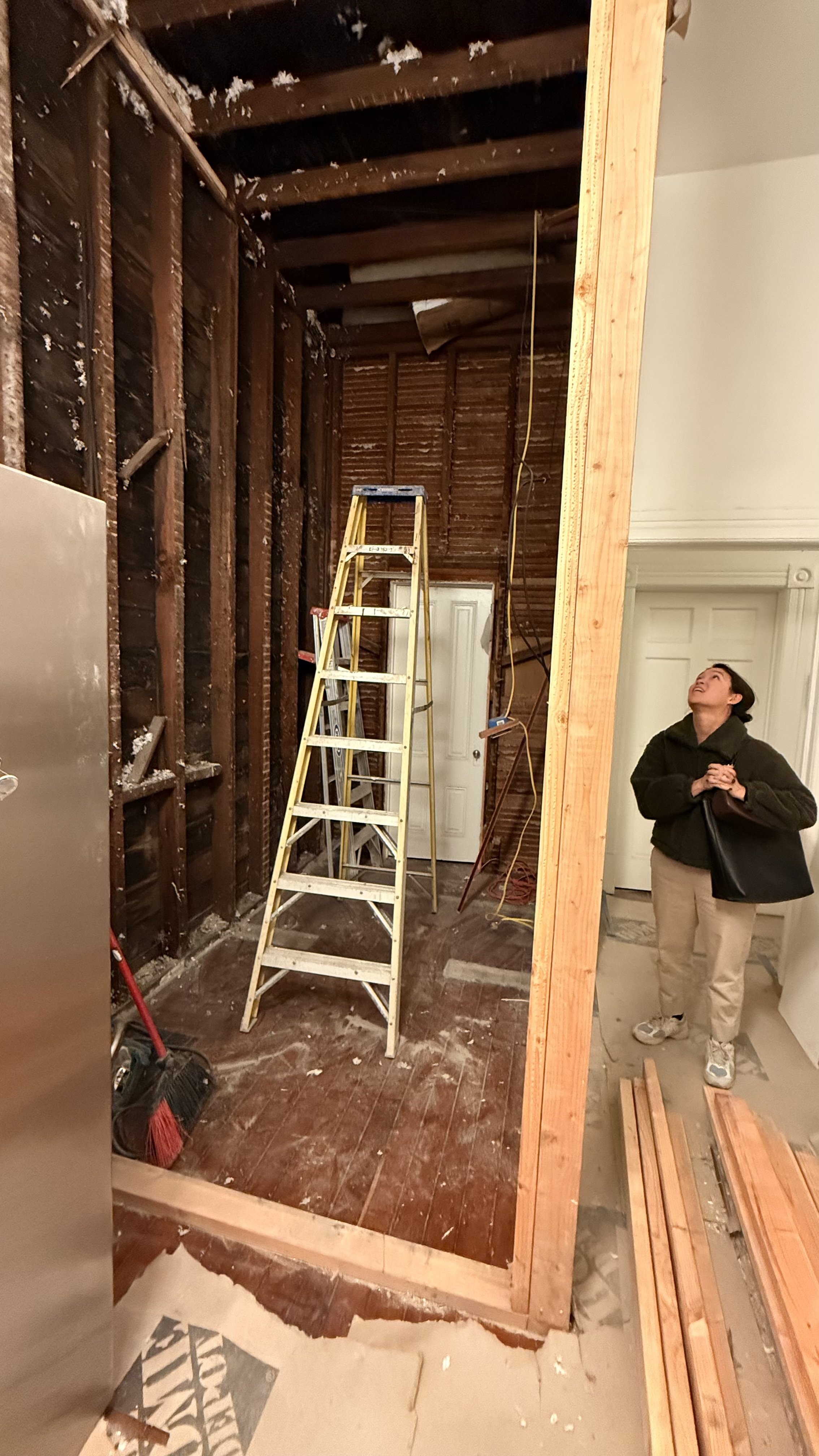 A woman standing inside a house under renovation, looking up at exposed wooden wall and ceiling framing. There is a ladder in the room, construction tools, and construction materials visible.