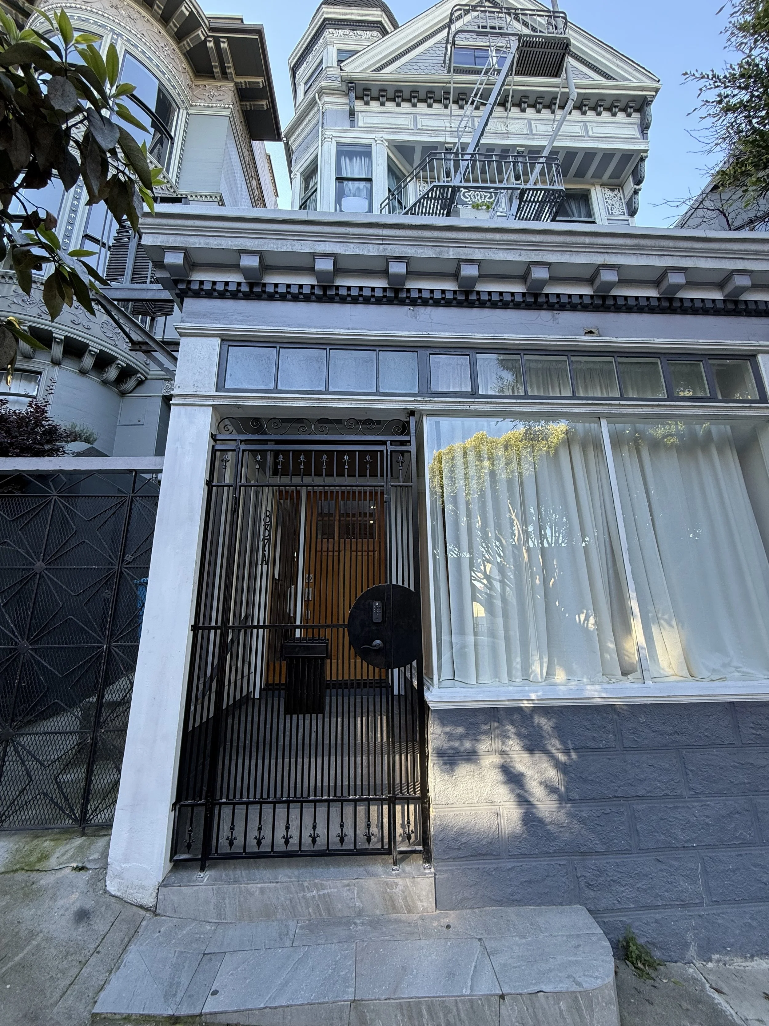 Facade of a Victorian-style house with a gated entry, large window with curtains, and a fire escape on the upper floors.