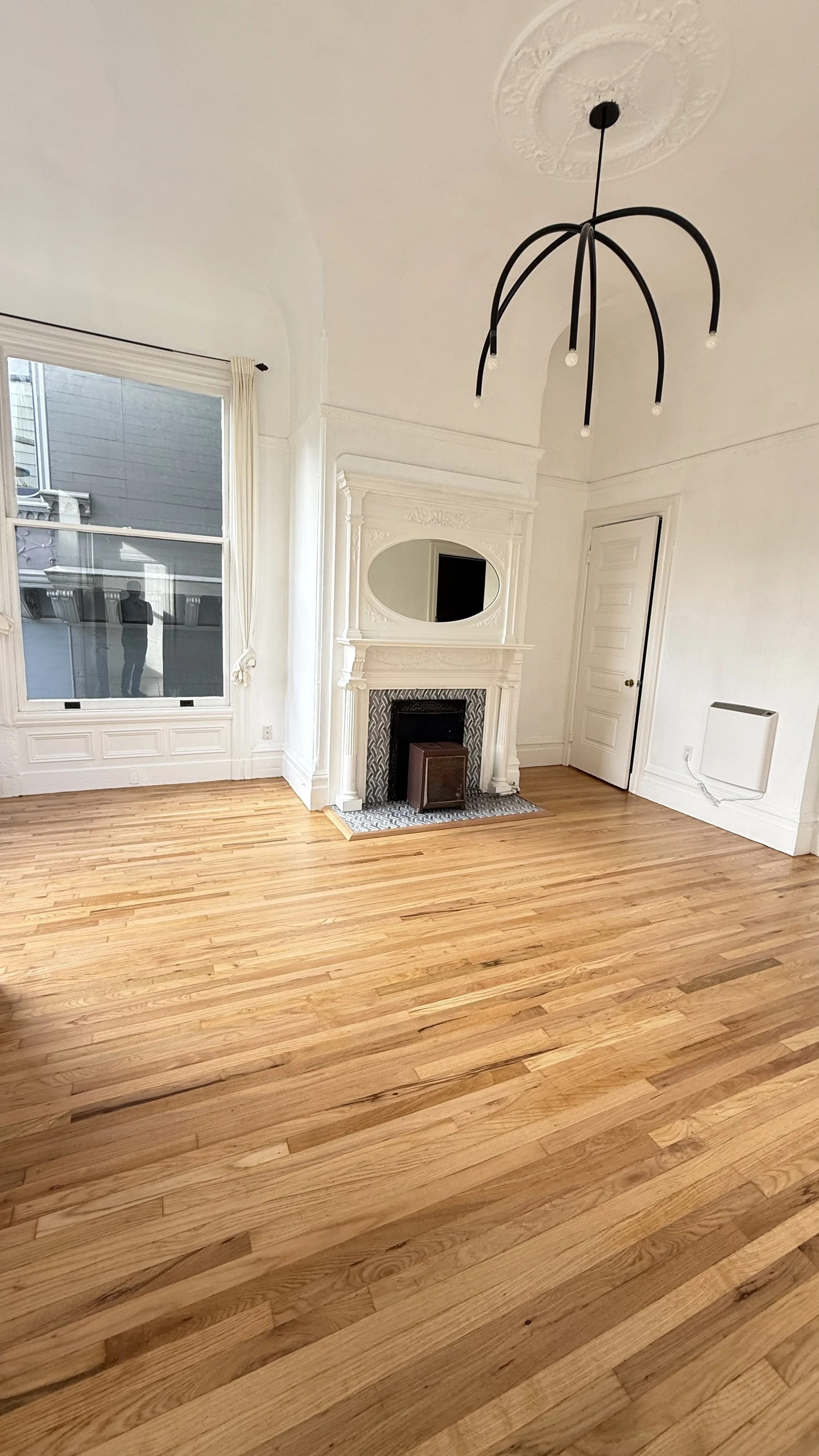 Empty room with hardwood floor, white walls, vintage fireplace with mirror above, large window, modern black chandelier, and a closed white door.