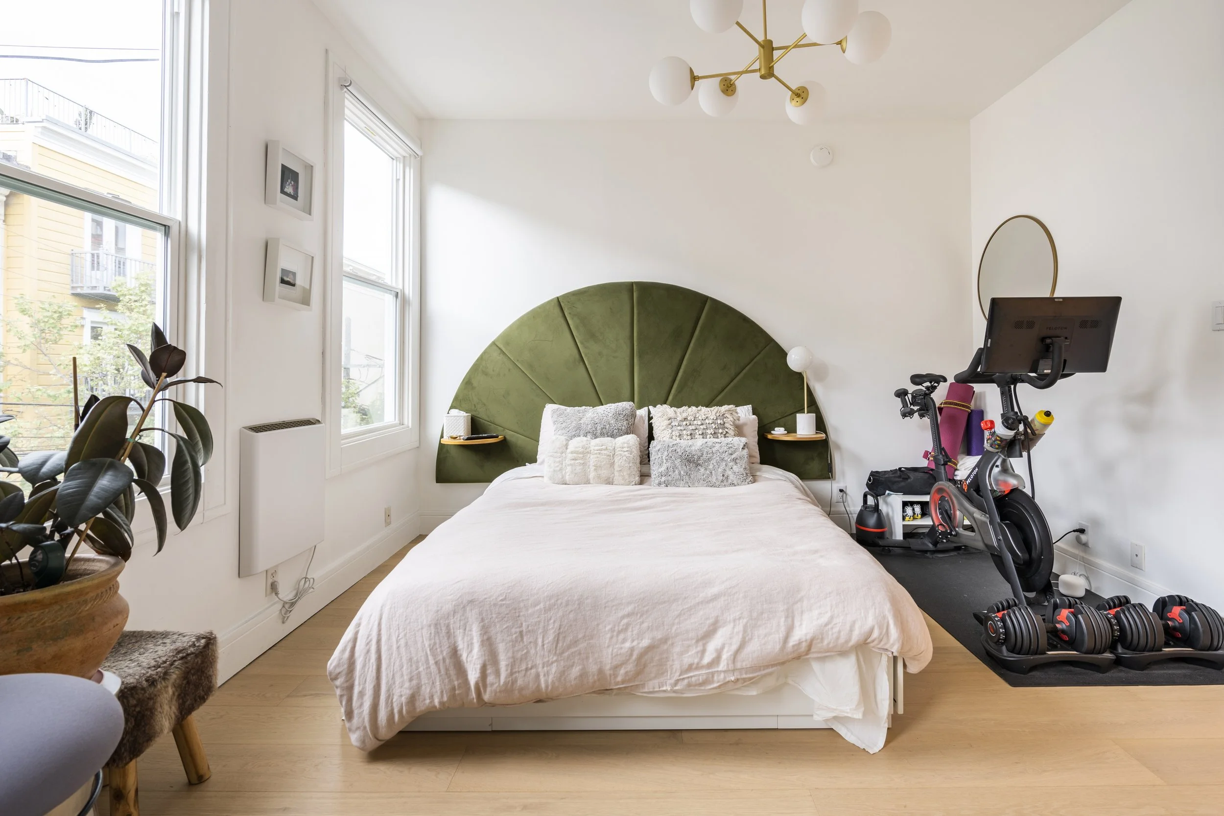 A bedroom with white walls and natural light from two windows, a green semi-circular headboard, a bed with multiple pillows, a wooden side table, exercise equipment including a stationary bike and dumbbells, and decorative items like framed photos and a mirror.