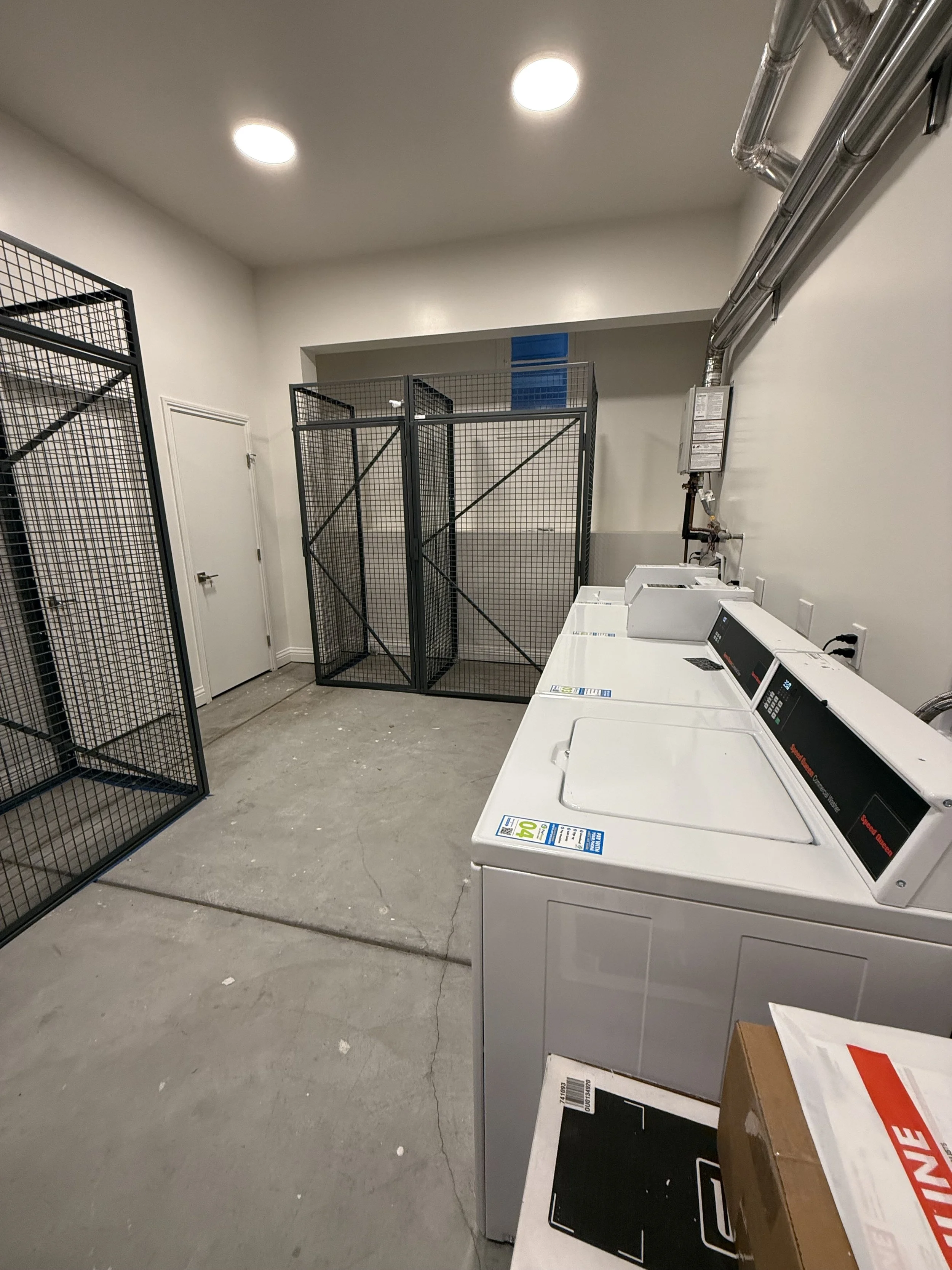 A laundry room with three white washing machines, a cardboard box, and black metal pet cages against the wall.