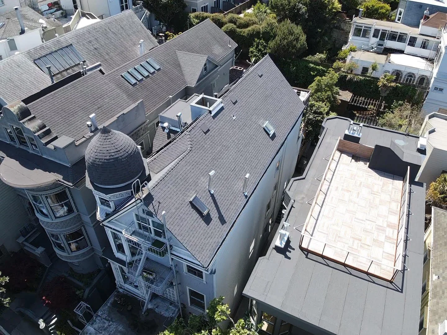 An aerial view of multiple rooftops of residential buildings in a densely populated neighborhood, showing various roof styles, skylights, chimneys, trees, and balconies.