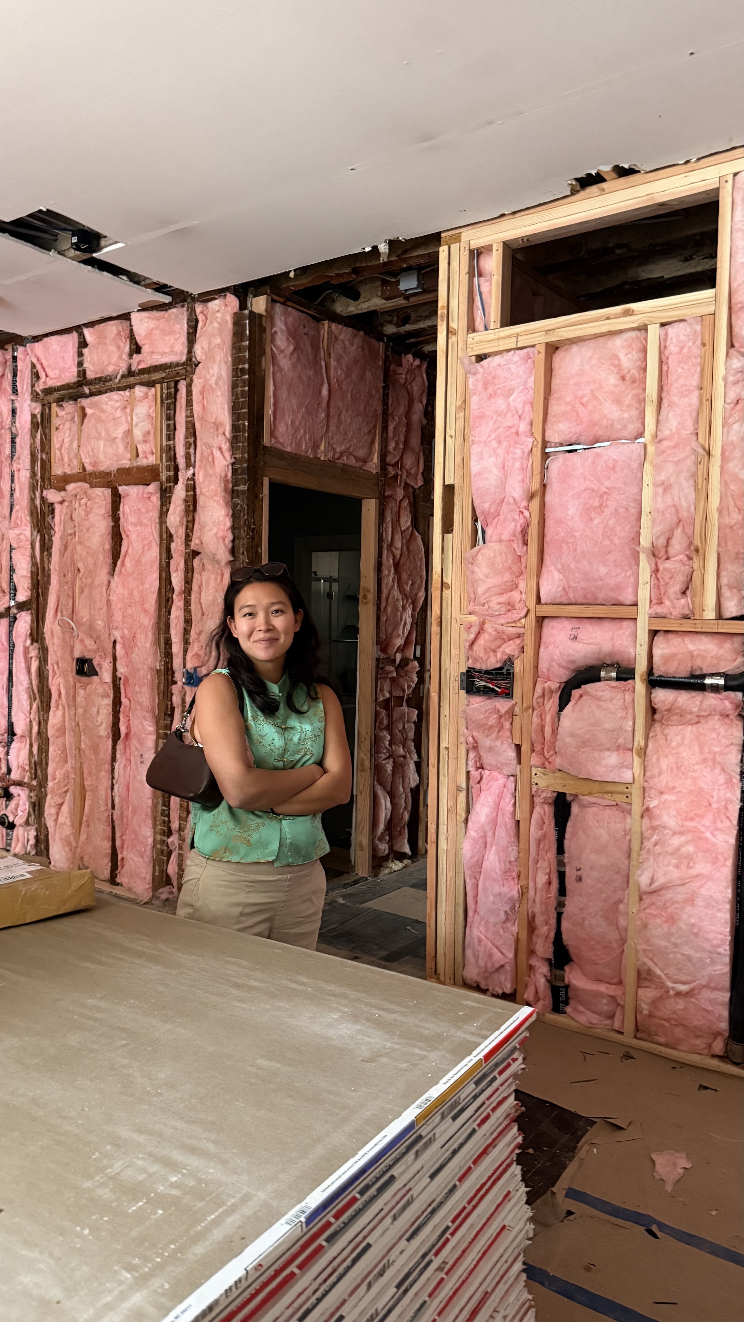A woman standing inside a house under renovation, surrounded by unfinished walls with pink insulation and wooden framing.