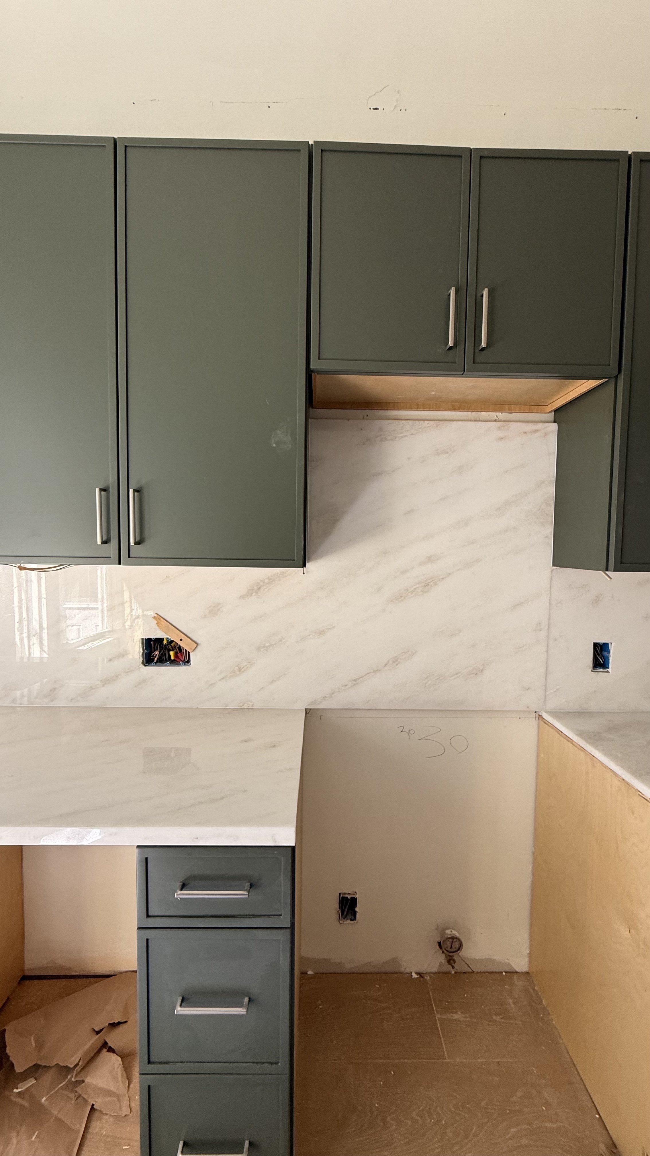 Kitchen under construction with green upper cabinets, a marble backsplash, and a dark green lower cabinet with drawers.