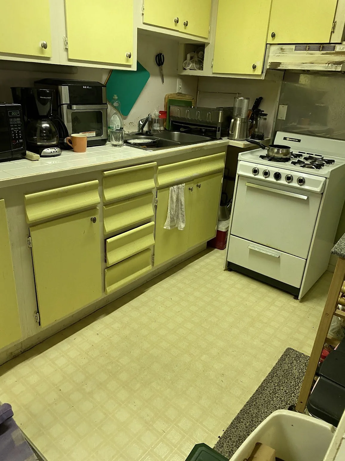 A small, cluttered kitchen with yellow cabinets, a white stove, and various appliances on the countertop, including a coffee maker and microwave.