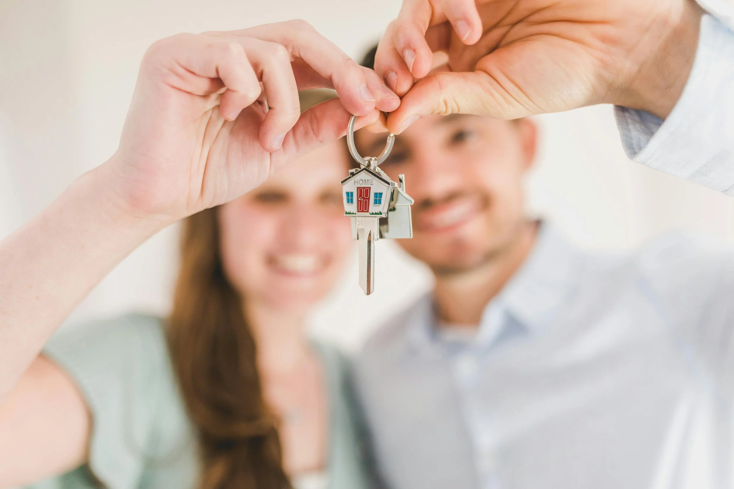 Couple holding up a house-shaped keychain, smiling at the camera, with their faces blurred in the background.