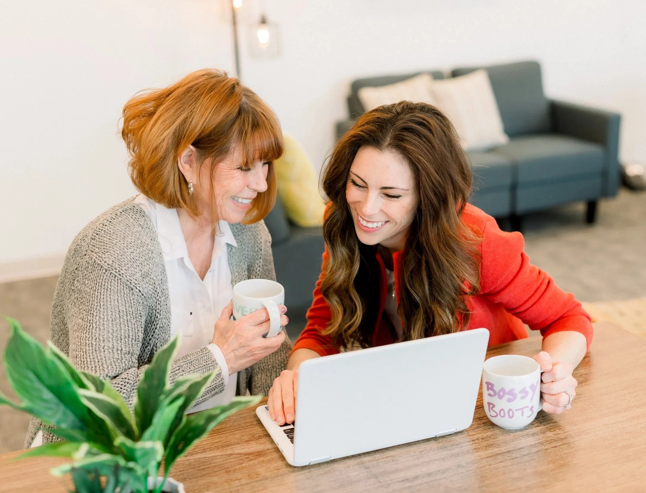 Two women sitting at a wooden table, looking at a laptop and smiling, with coffee mugs in hand, in a cozy living room with a couch and a plant in the foreground.