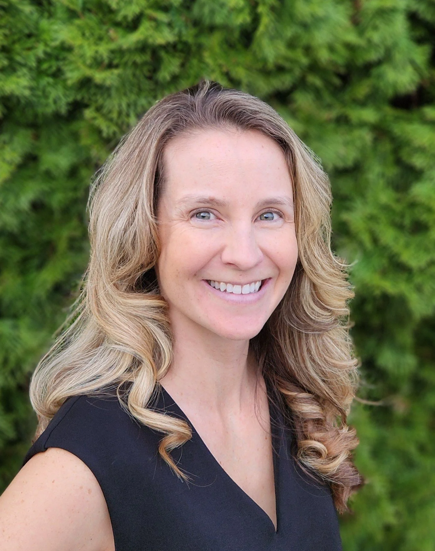A woman with long, curly blonde hair is smiling and standing outdoors in front of green foliage.