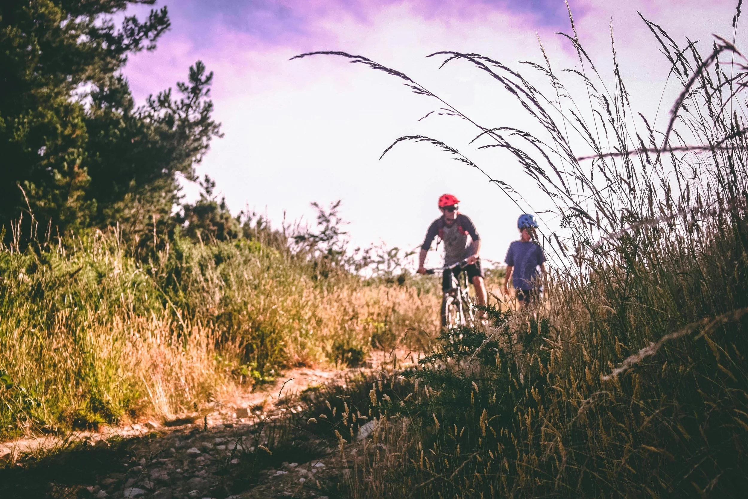 Two people riding bikes on a trail through a grassy and wooded area, wearing helmets, with the sky in the background.