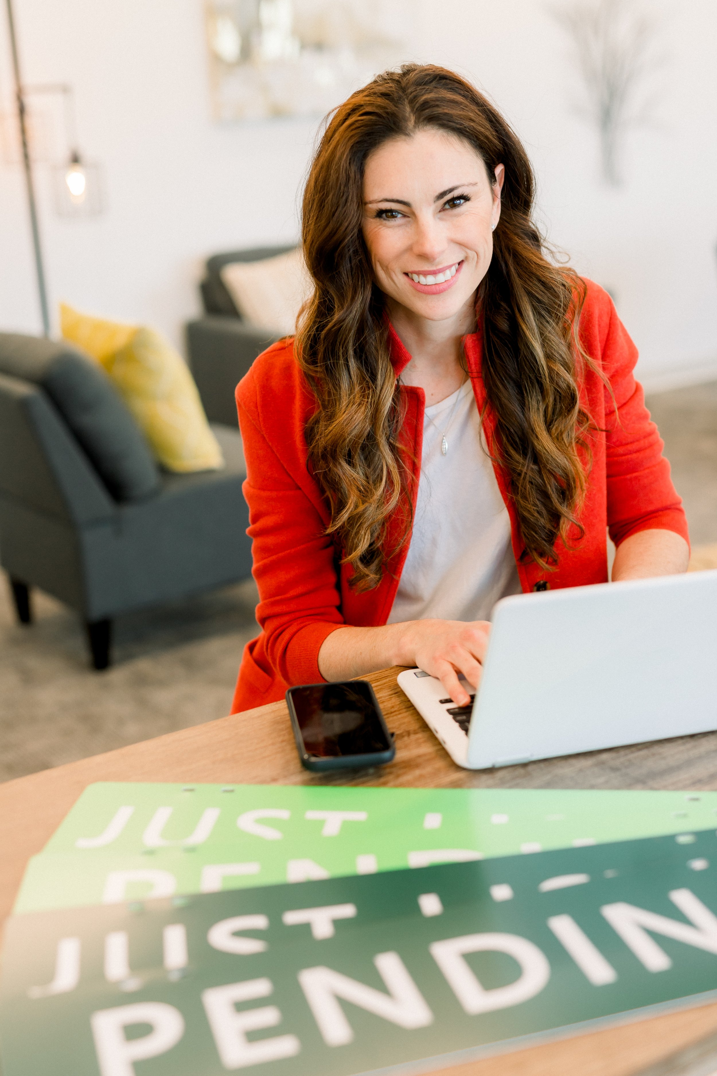 A woman with long brown hair, wearing a red jacket and white top, smiling while sitting at a desk with a laptop and smartphone. There are green and white signs on the desk that read 'Just Be PENDING.'