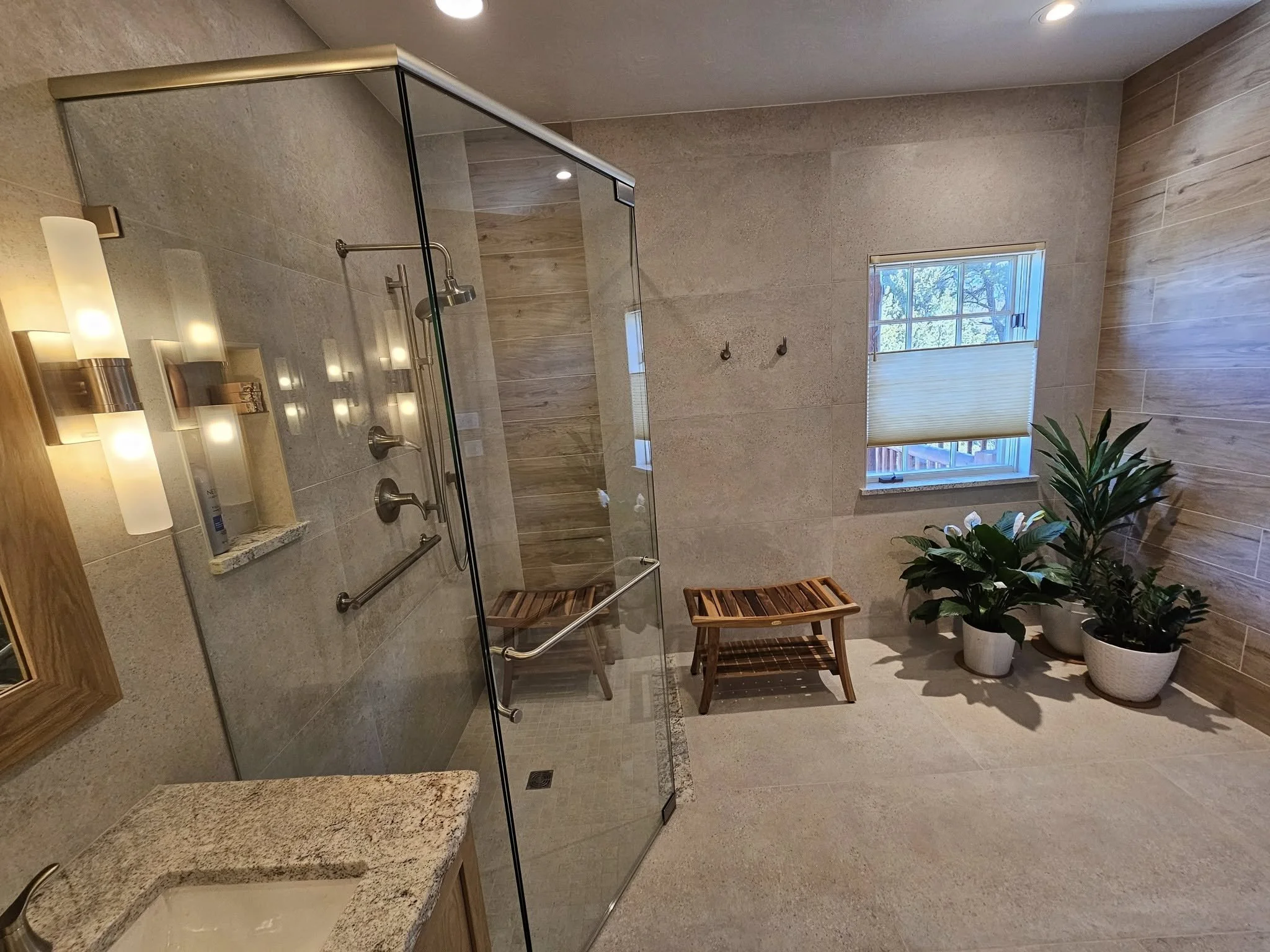 Bathroom with a glass shower, wooden bench, and potted plants near window with blinds.