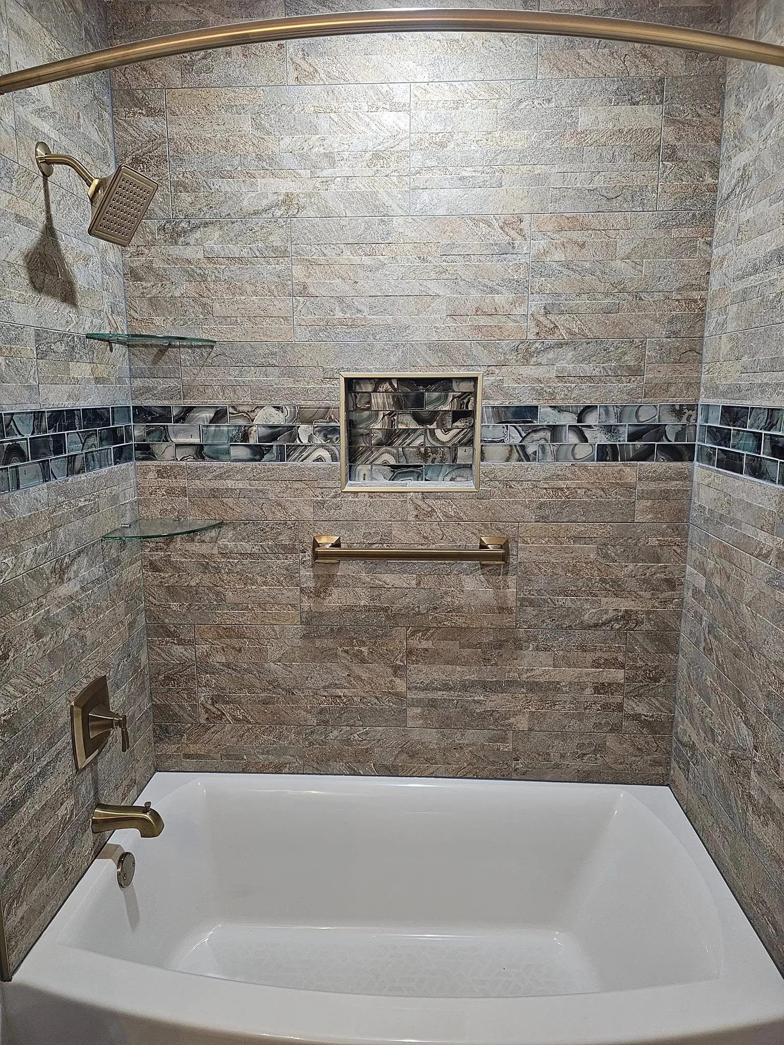 A tiled bathroom shower with a bathtub, glass shelves, a metallic showerhead, a grab bar, a recessed shelf, and a wall-mounted faucet.