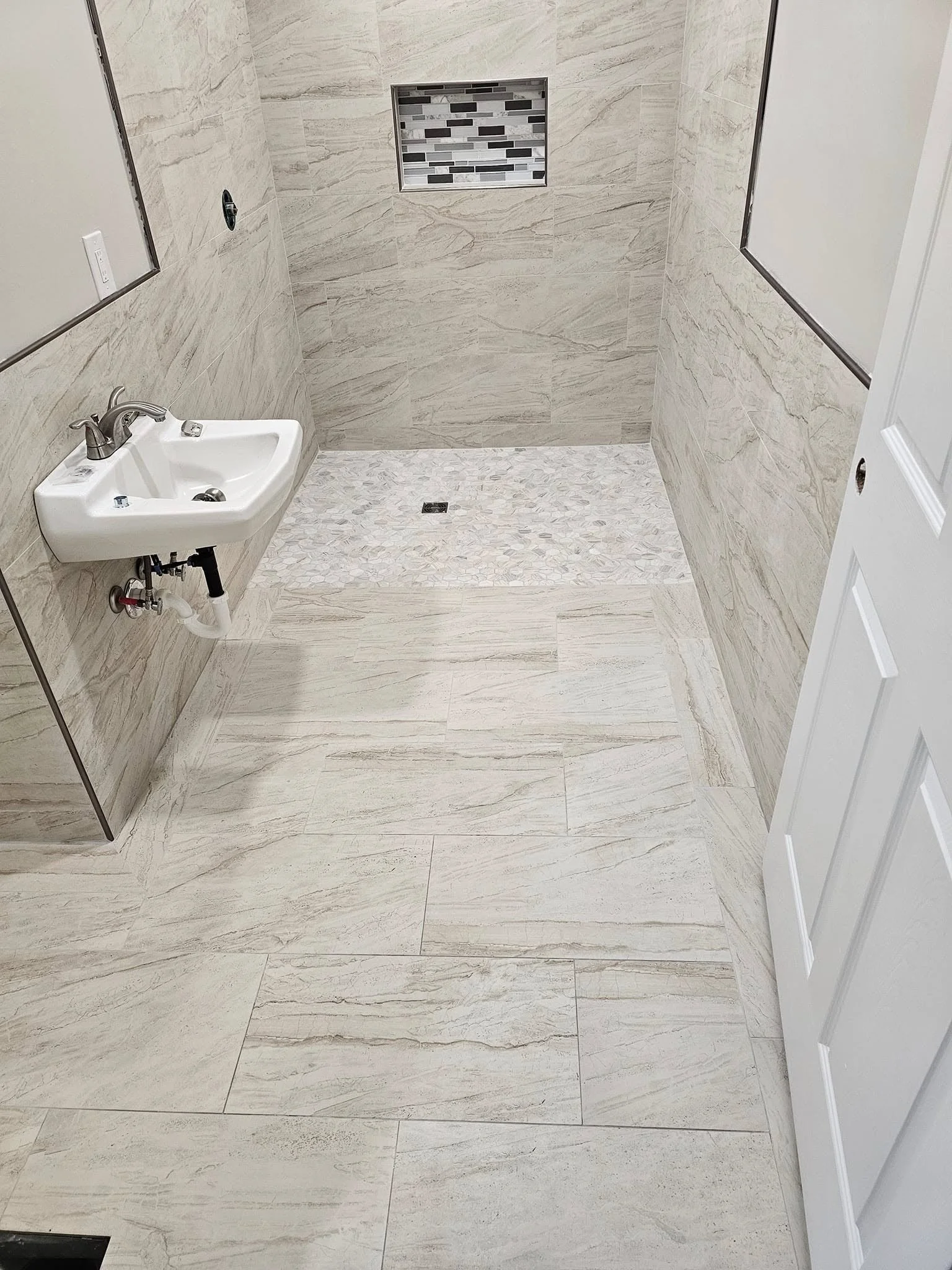 Bathroom with beige marble tiles, small white sink on the left, mirror frames on the walls, and a shower area with tile mosaic and niche in the far wall.
