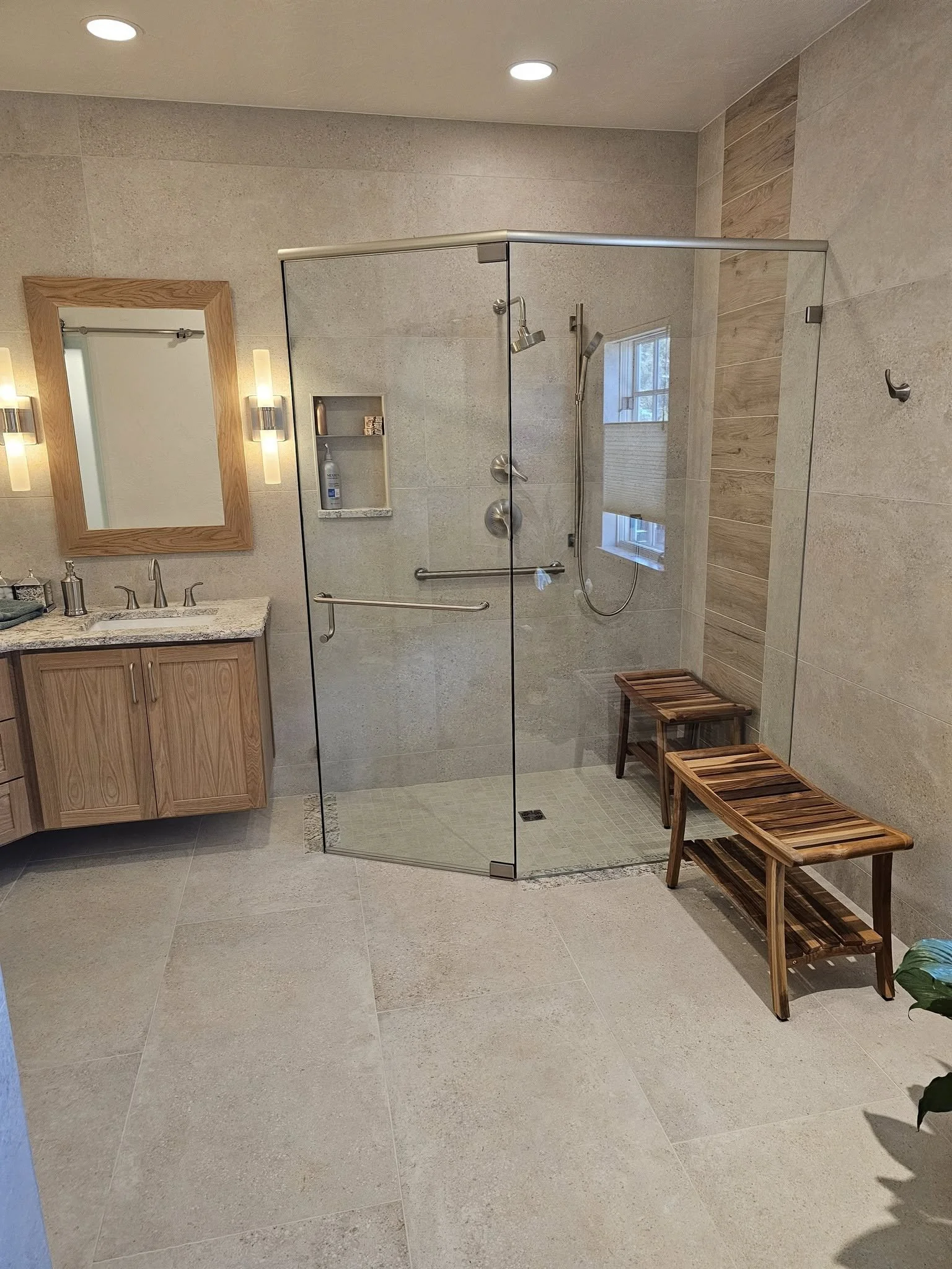 Modern bathroom with walk-in glass shower, wooden vanity with mirror, beige tiled floor and walls, two wooden benches, and ceiling lights.