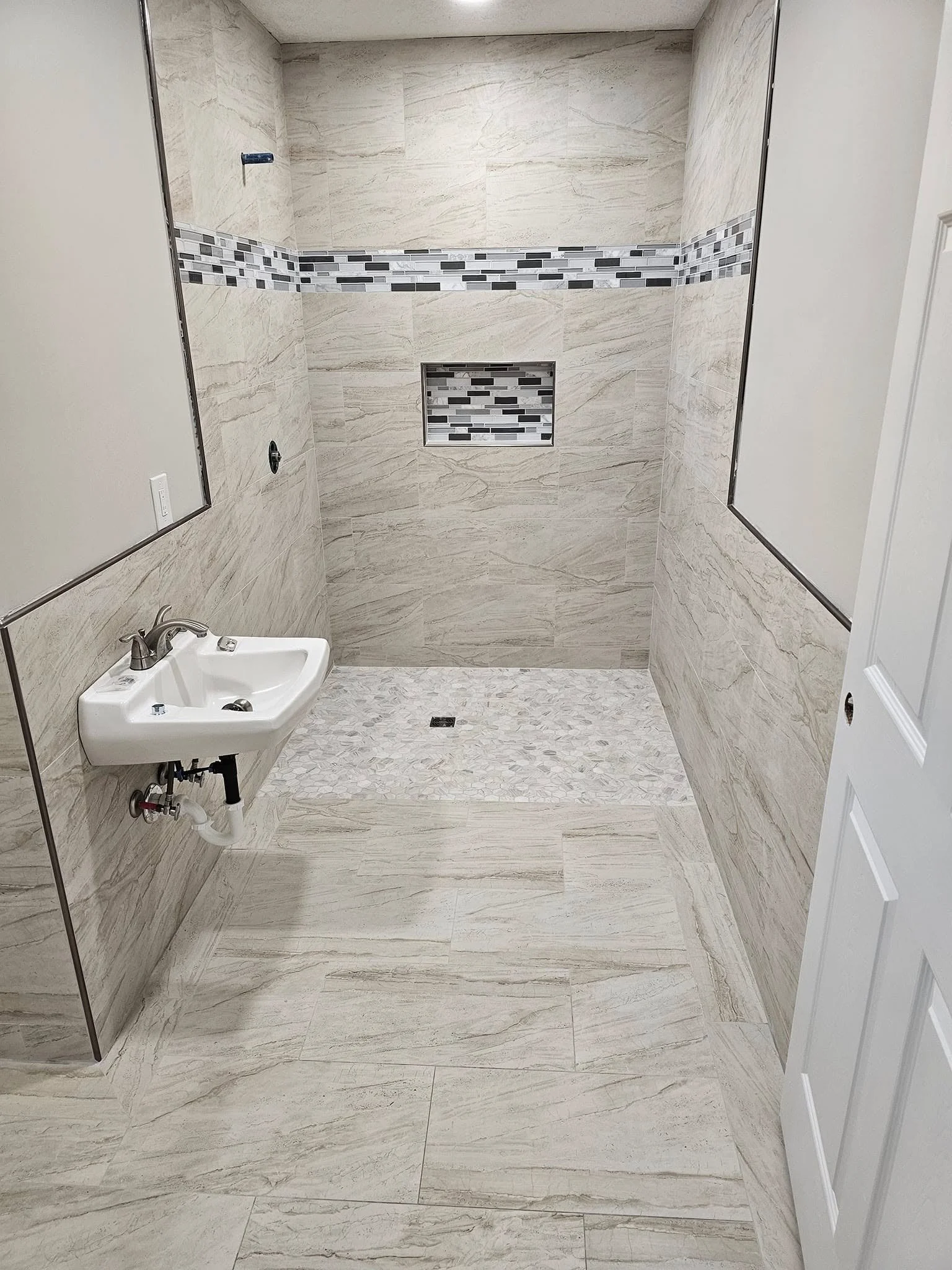 Empty bathroom with beige marble tile walls and floor, a small white wall-mounted sink on the left, and a large walk-in shower with a built-in niche and decorative tile accents on the far wall.
