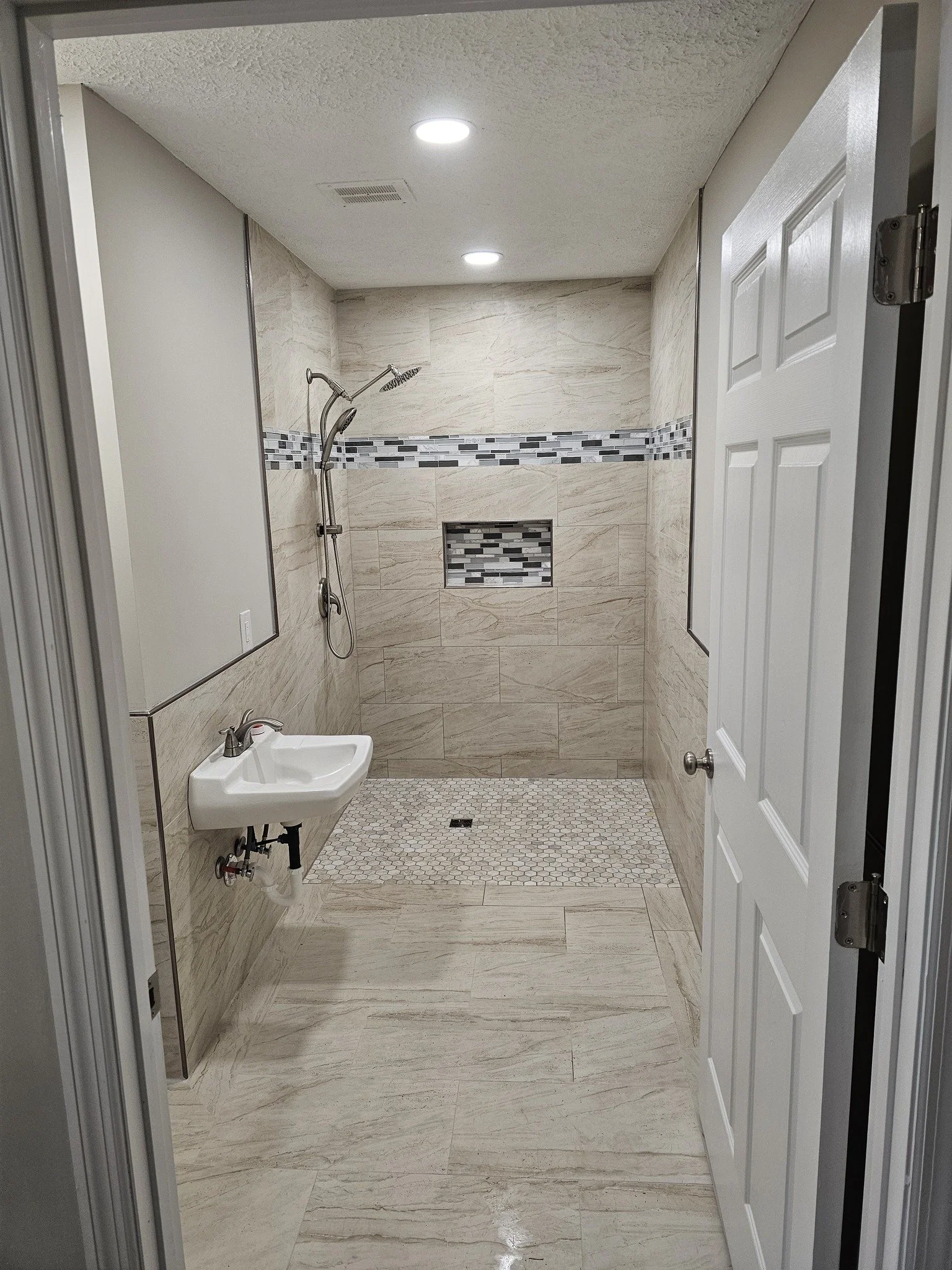 A modern bathroom with a walk-in shower, small hexagonal floor tiles, beige wall tiles with a decorative horizontal gray and black mosaic strip, a small wall-mounted sink, and a dirty white door inside a white frame.