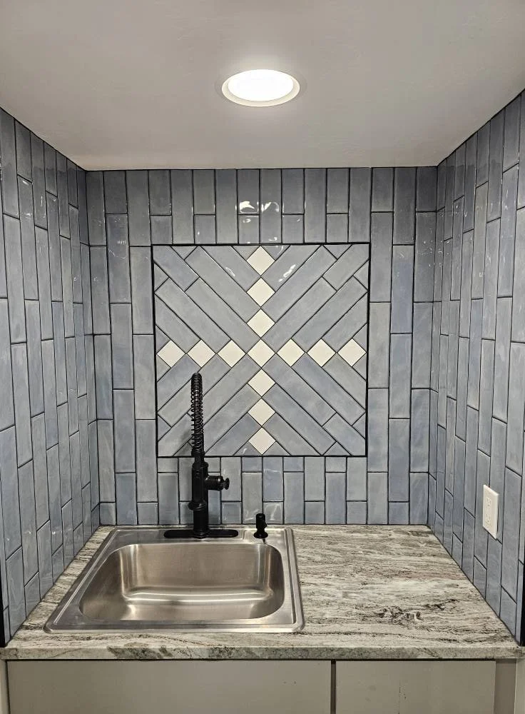 Kitchen area with a stainless steel sink, black faucet, and gray patterned tile backsplash.