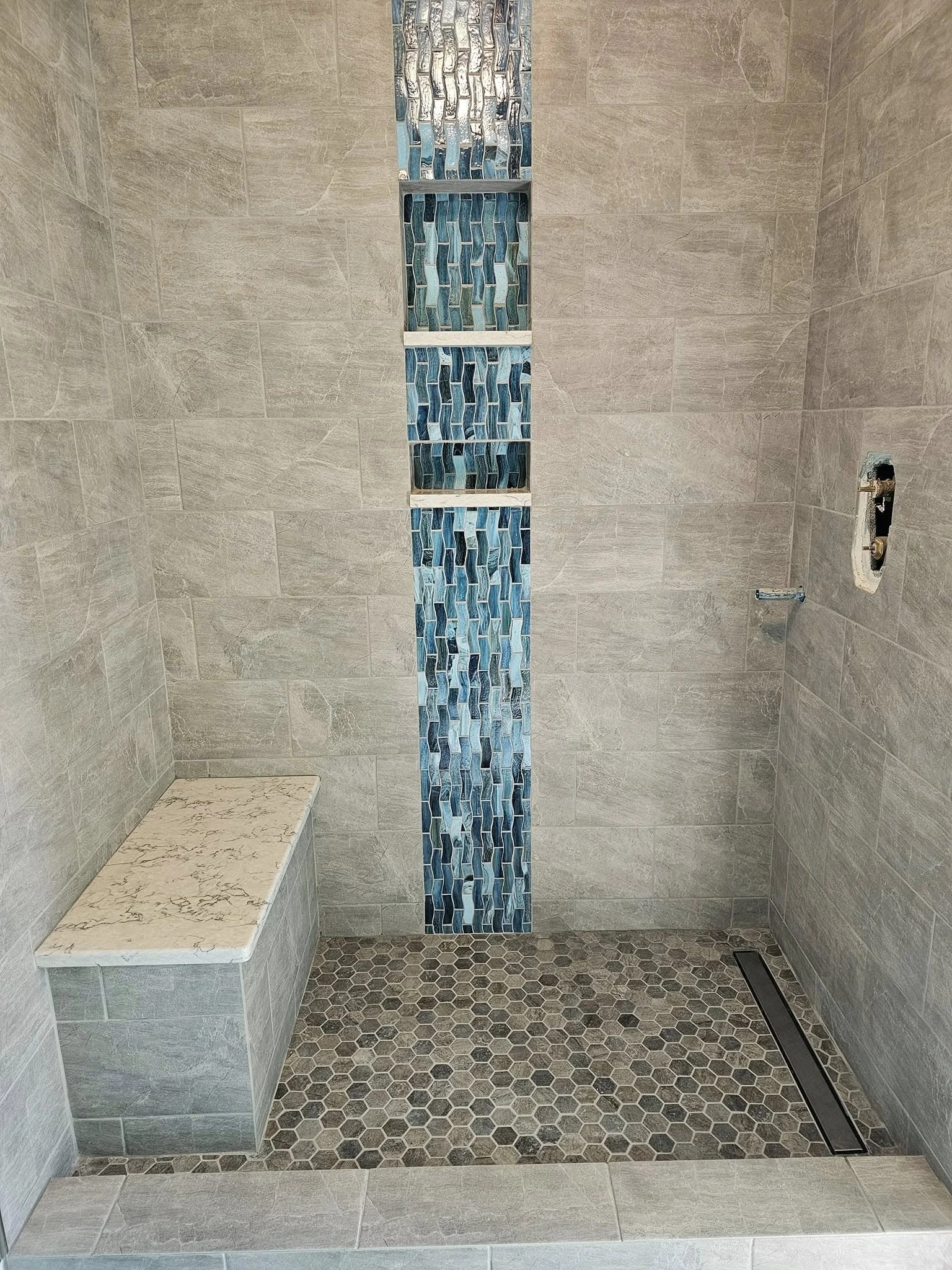 Empty bathroom shower with beige tile walls, a vertical strip of blue mosaic tiles, a marble seat on the left, and a floor drain along the right side.
