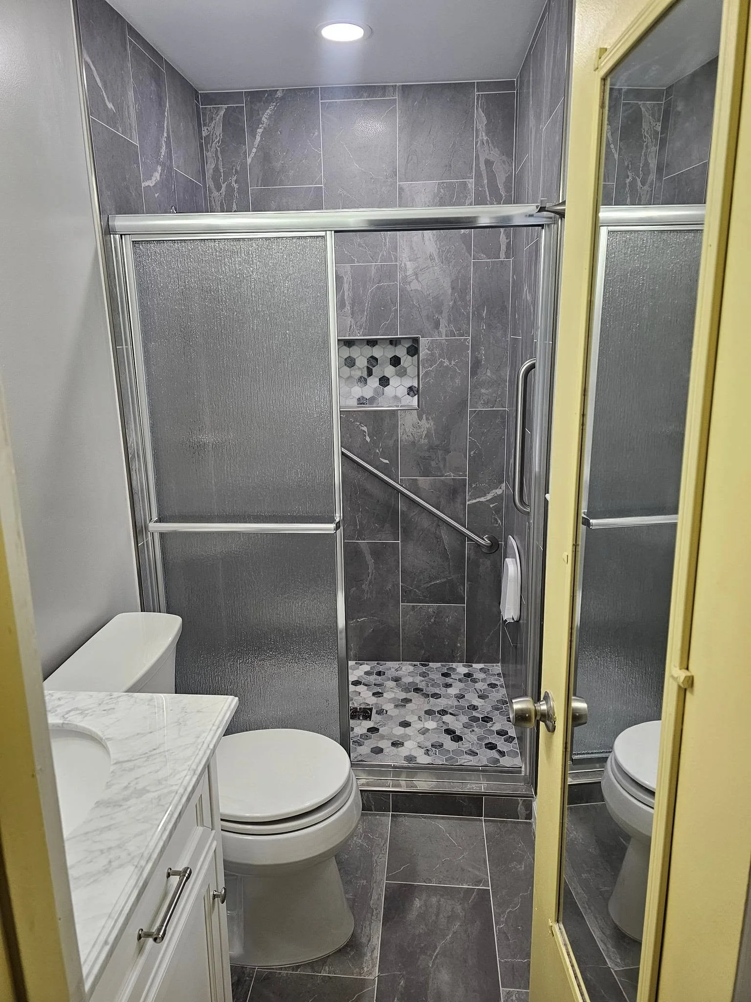 Small bathroom with a shower stall, toilet, and vanity with a marble countertop, yellow door frame, and gray tiled walls and floors.
