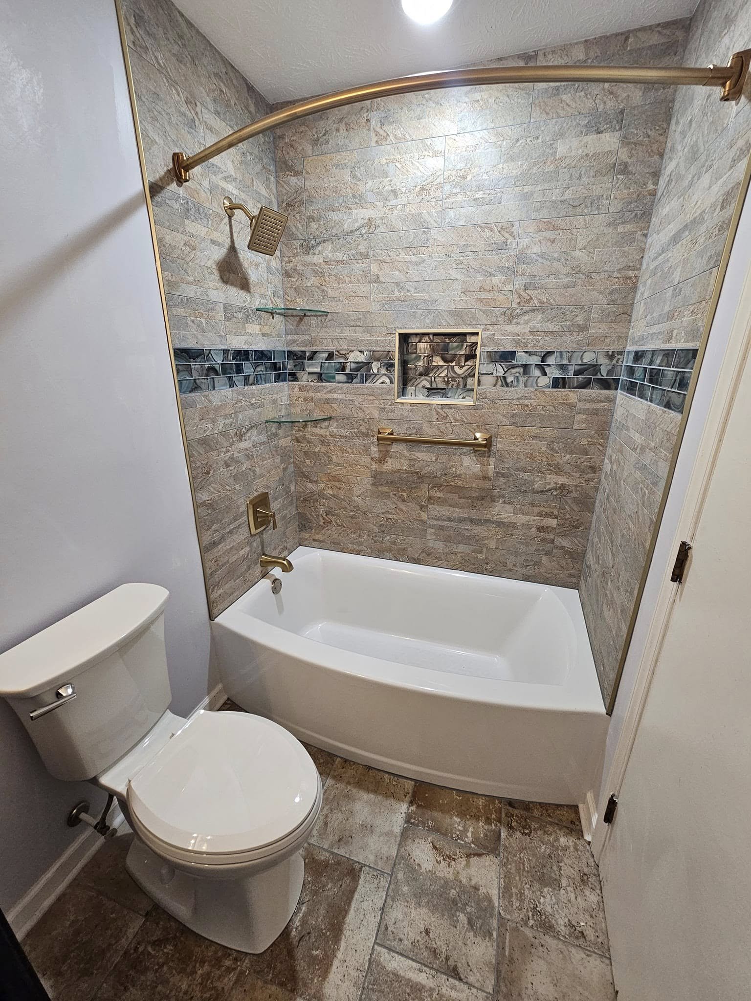 Bathroom with a bathtub and a toilet. The shower area has beige and gray stone tile walls with a decorative strip of black and gray tiles. There are two glass shelves, a built-in niche, a showerhead, a grab bar, and a faucet in the shower area.