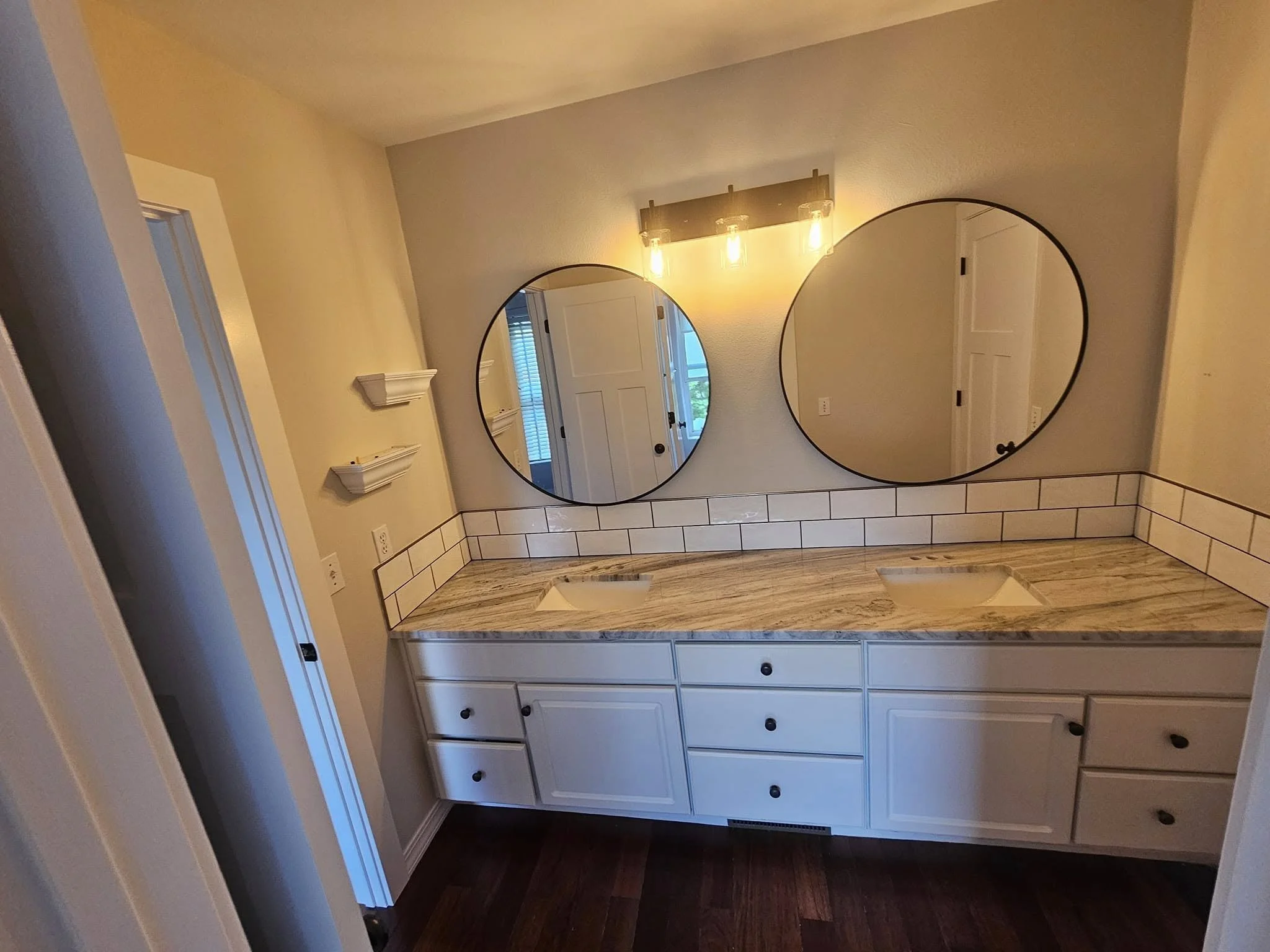 Bathroom vanity with a granite countertop, two sinks, and two round mirrors above it, beige walls, and wooden flooring.
