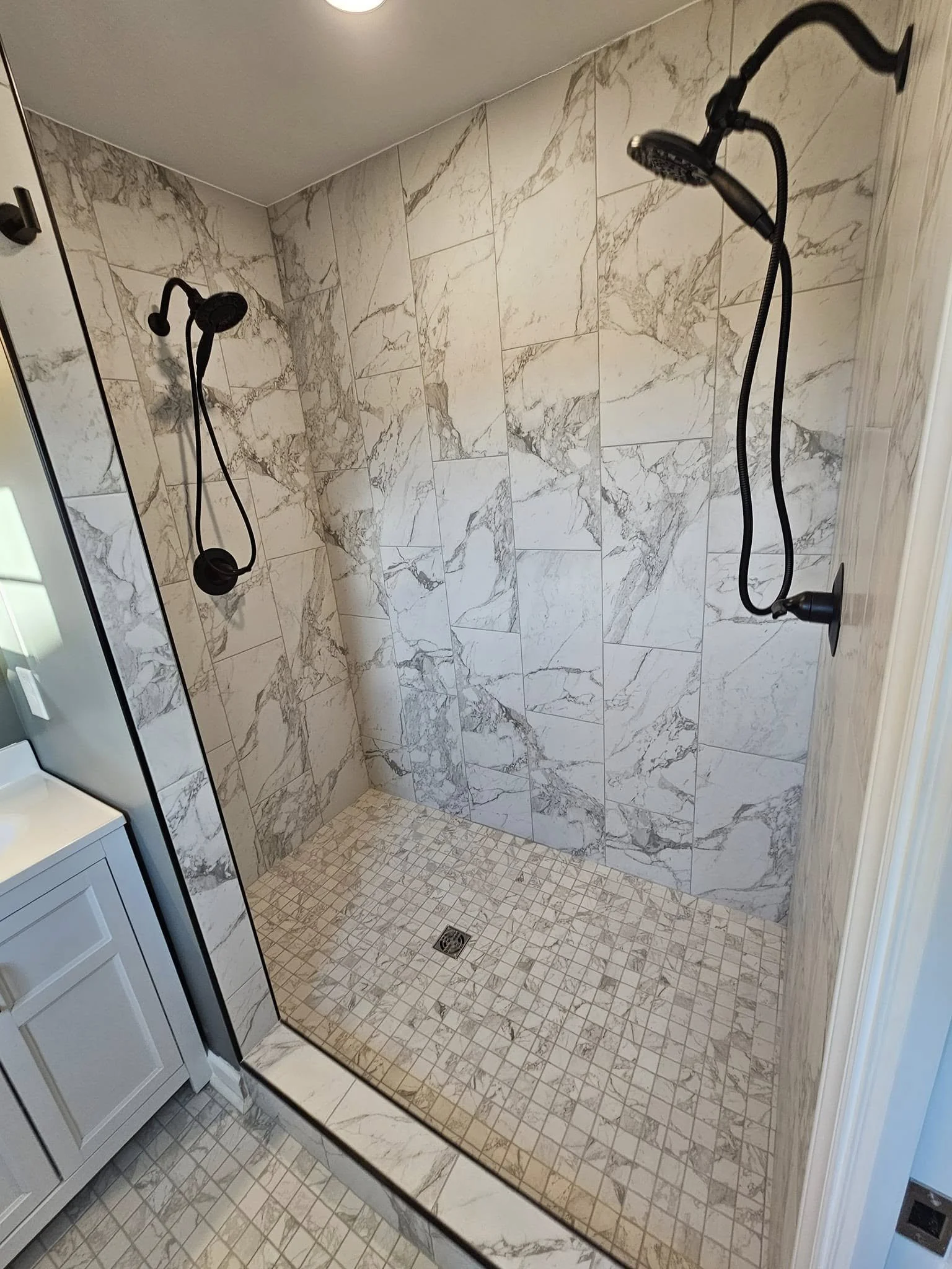 A modern walk-in shower with marble tiles on the walls and small mosaic tiles on the floor, featuring two black handheld shower heads mounted on the wall.