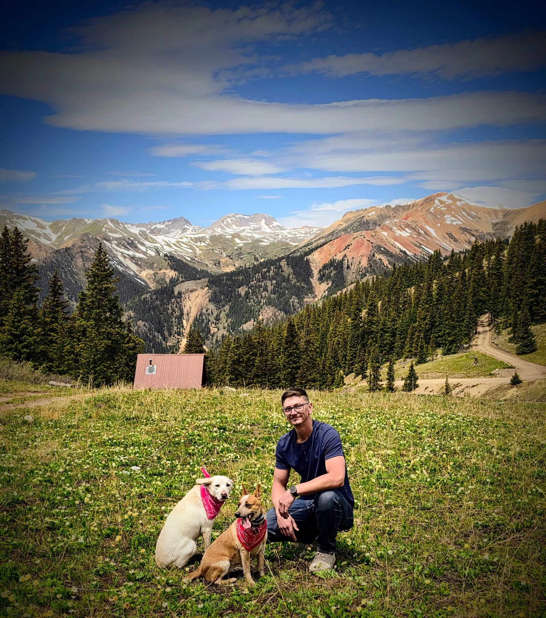 A man kneeling in a grassy field with three dogs, one wearing a pink bandana, in a mountainous landscape with a partly cloudy sky.