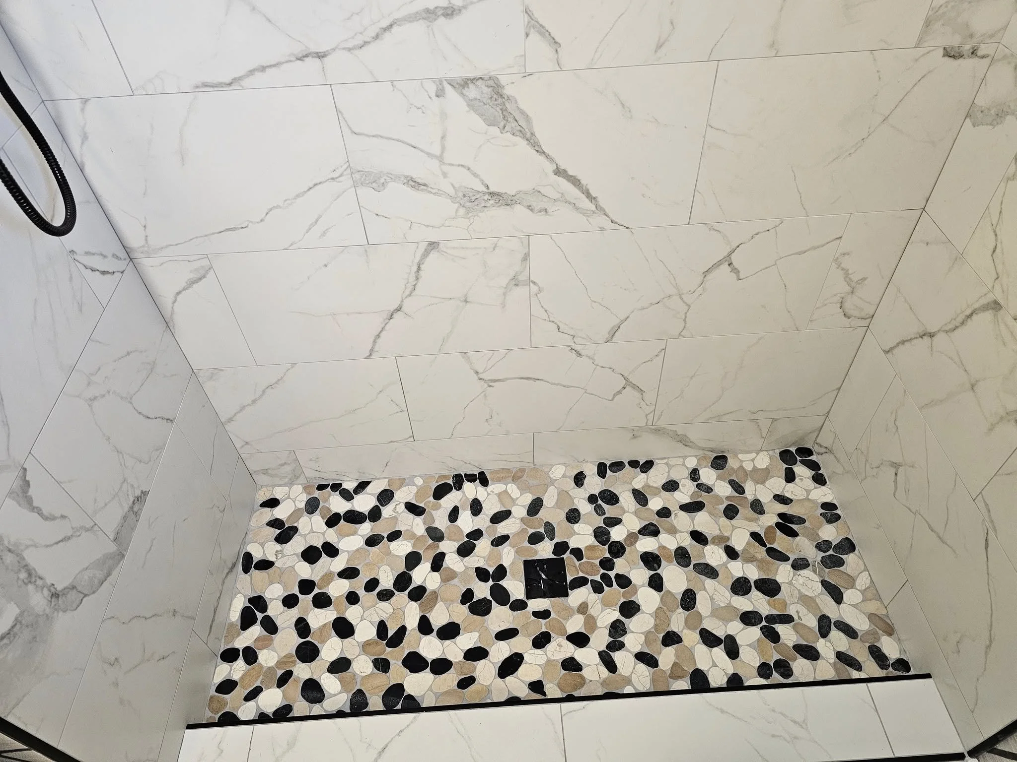 Inside a shower with large white marble tiles on the walls and a small pebble tile floor with black, white, and beige stones, and a black drain in the center.