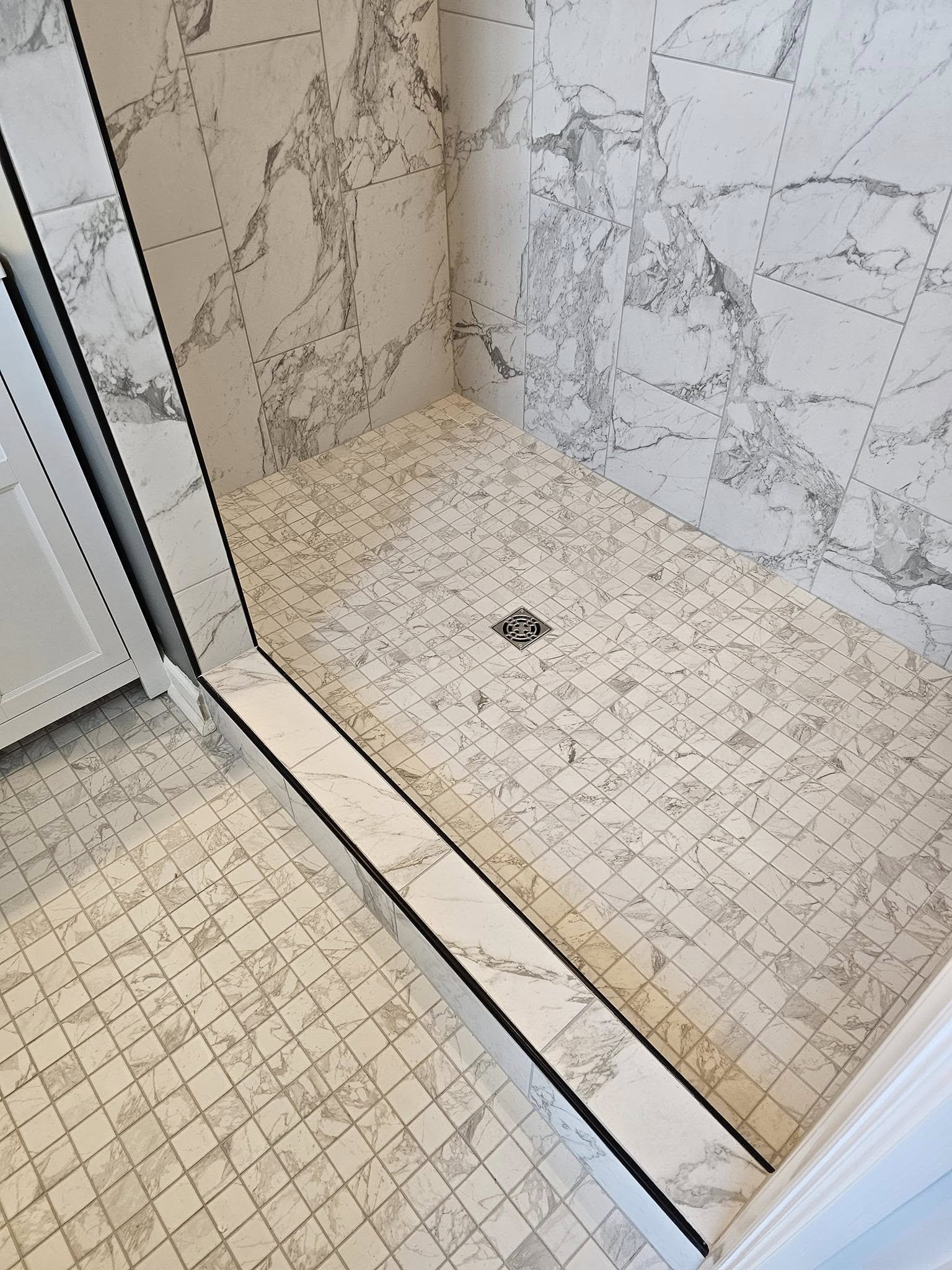 A shower area with marble wall tiles and beige small square floor tiles, featuring a drain in the center.