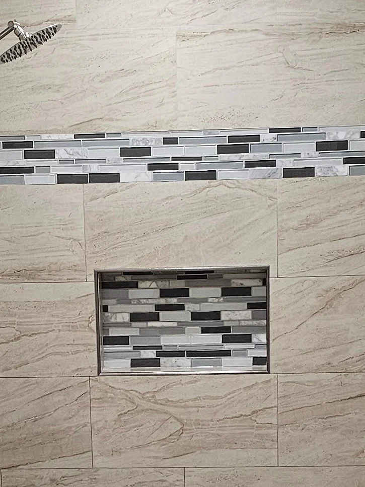 Close-up of a tiled shower wall with beige stone tiles, a horizontal mosaic strip, and a built-in rectangular niche with matching mosaic tiles.