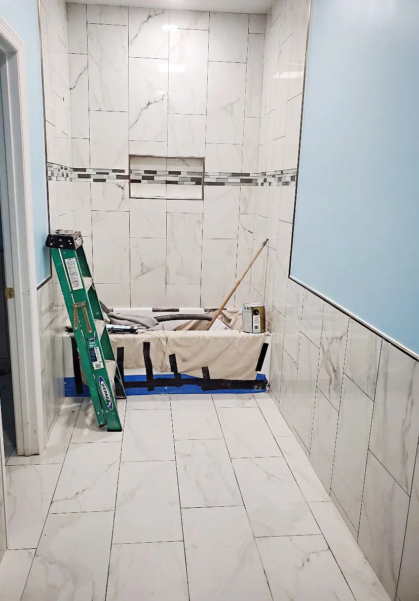 Bathroom with white marble tiling, partially tiled shower area under construction, a green ladder, a bucket, and construction tools.