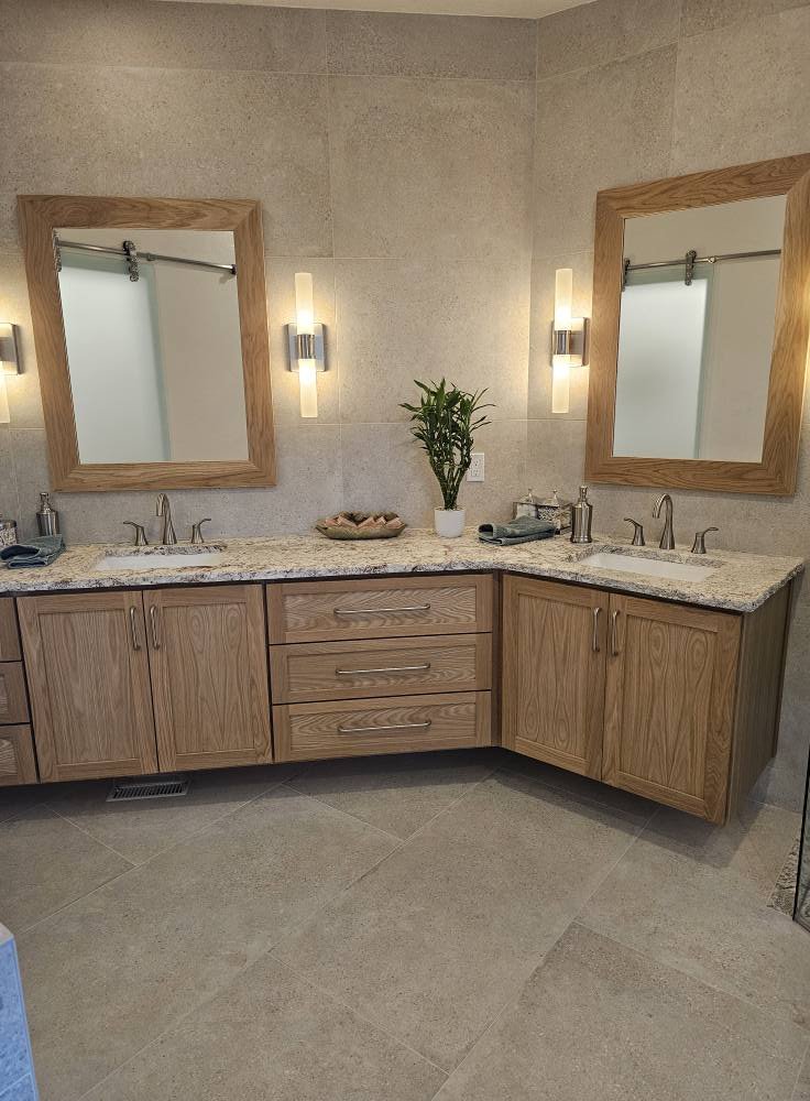 Double bathroom vanity with granite countertop, wooden cabinets, two mirrors, wall-mounted lights, and a green plant in the center.