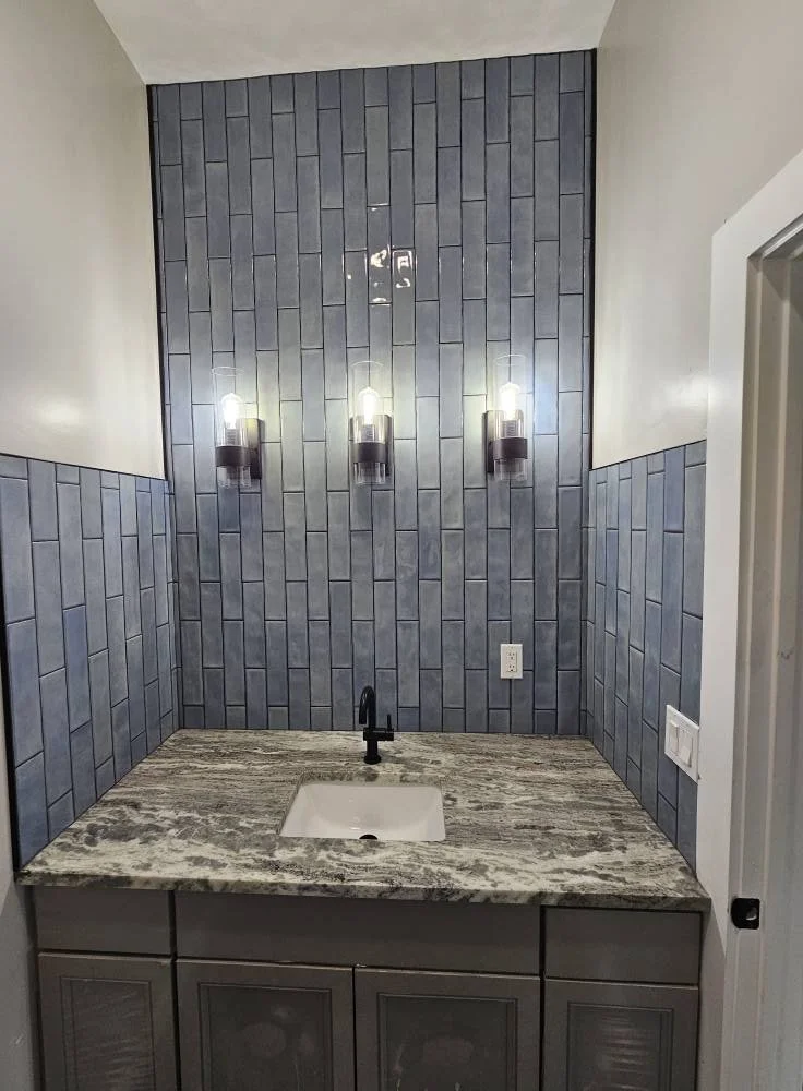 Bathroom vanity with a granite countertop, single sink, black faucet, blue tile backsplash, and three wall-mounted lights.